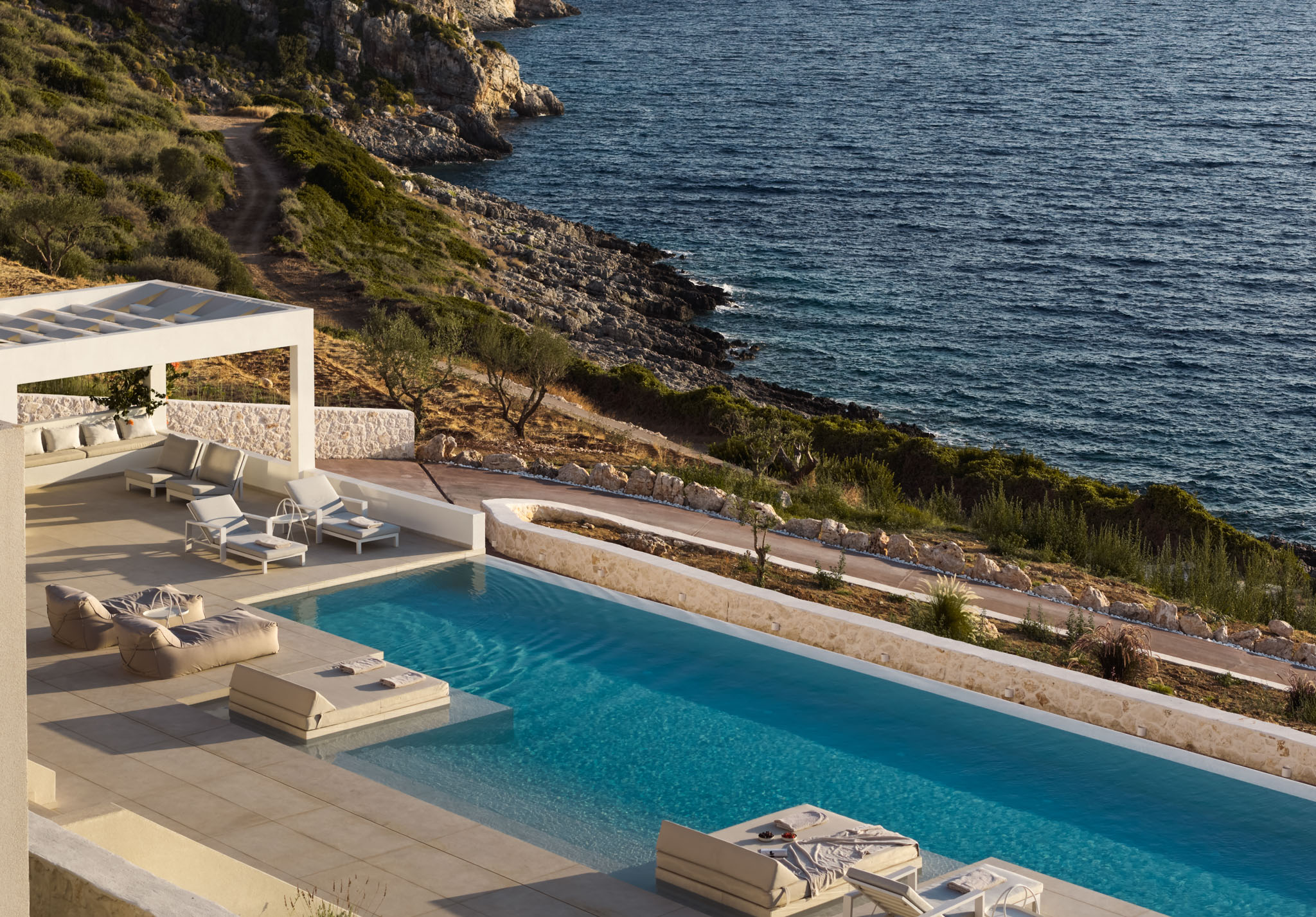 Elevated view of a long infinity pool and sun terrace with a white pergola lounge, overlooking a rocky, green coastline and deep blue sea.