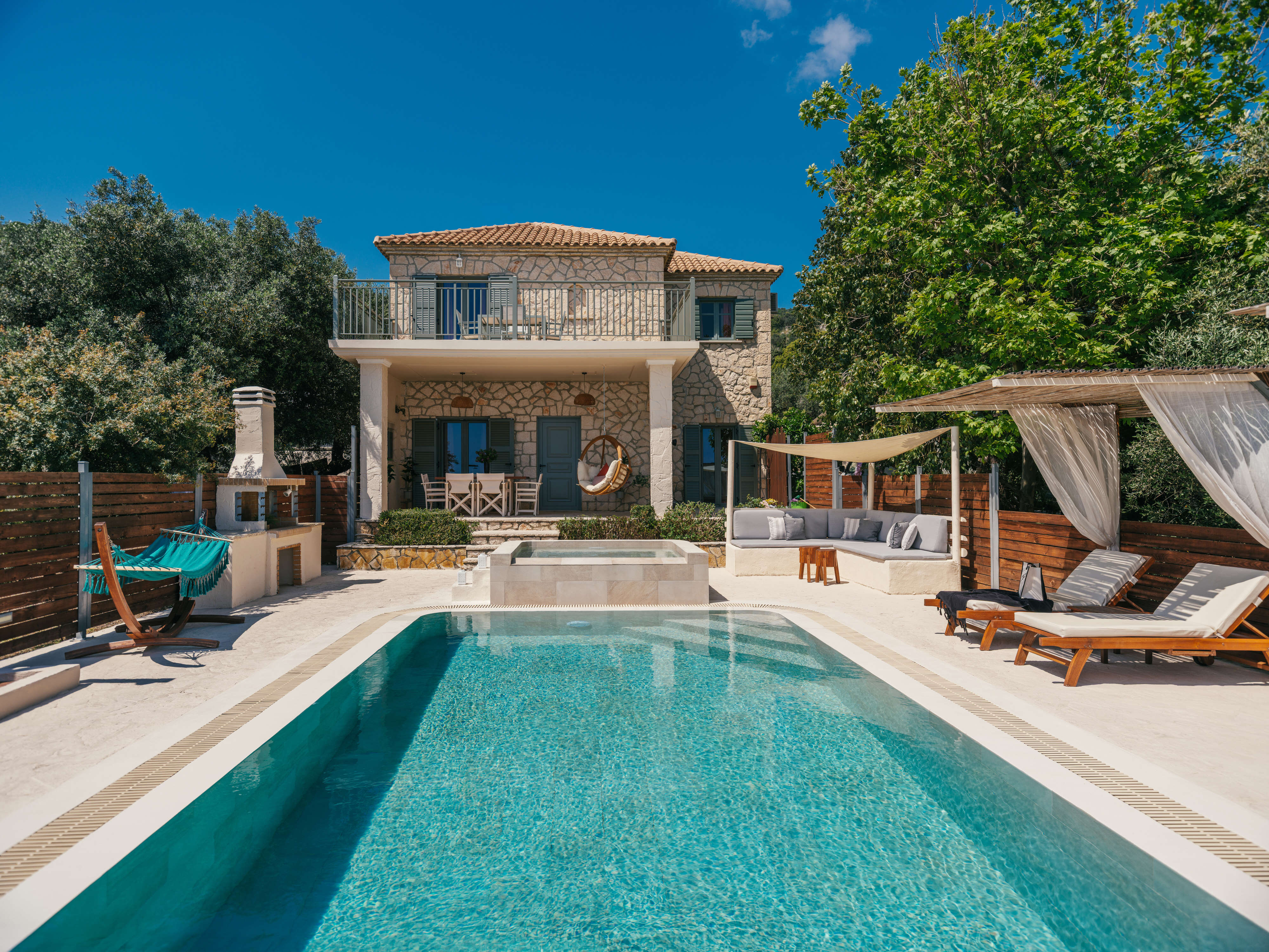 Front view of the two-storey stone villa with balcony and terrace, showing a turquoise hammock, hot tub, and wooden loungers around the rectangular pool.