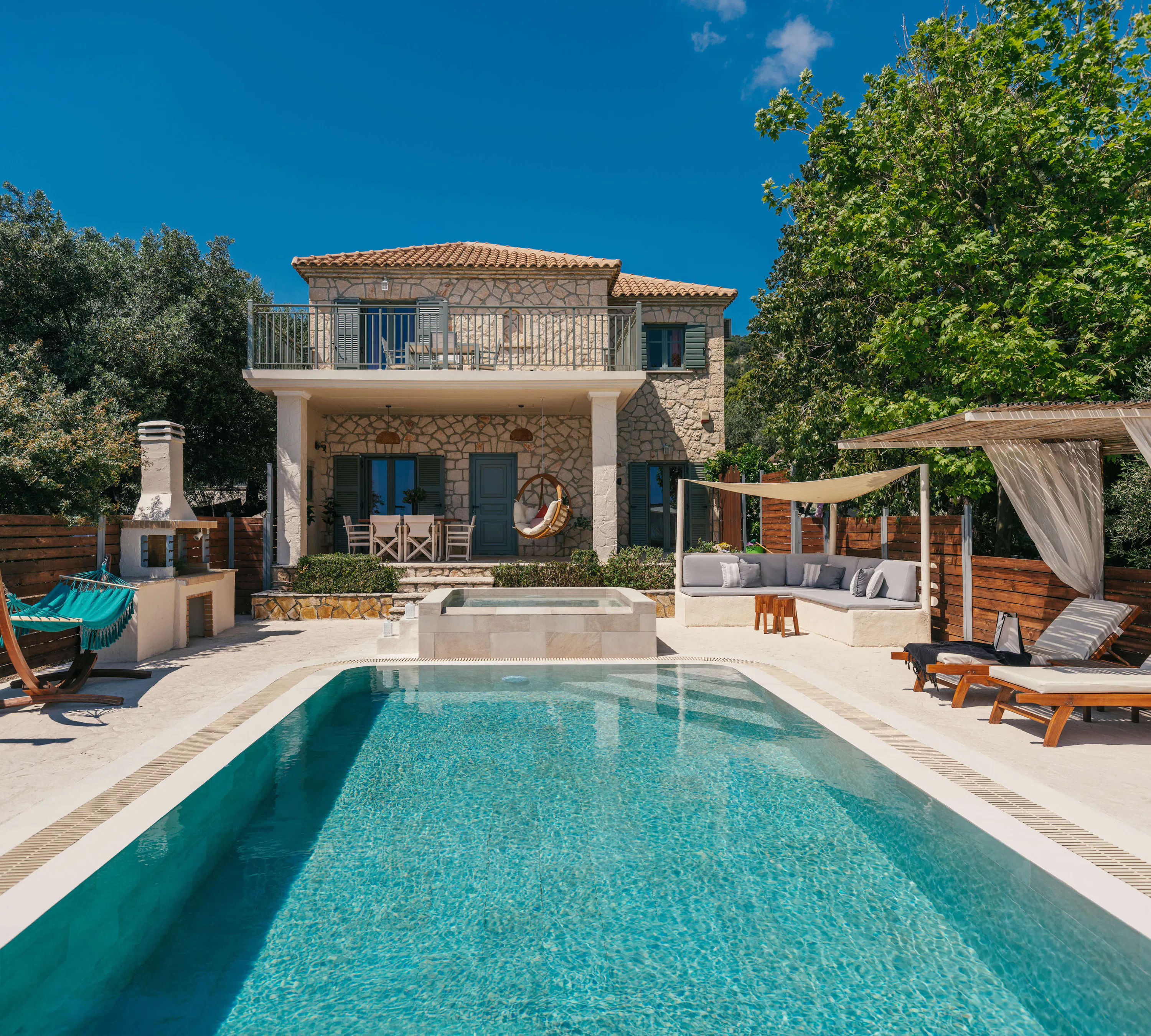 Front view of the two-storey stone villa with balcony and terrace, showing a turquoise hammock, hot tub, and wooden loungers around the rectangular pool.
