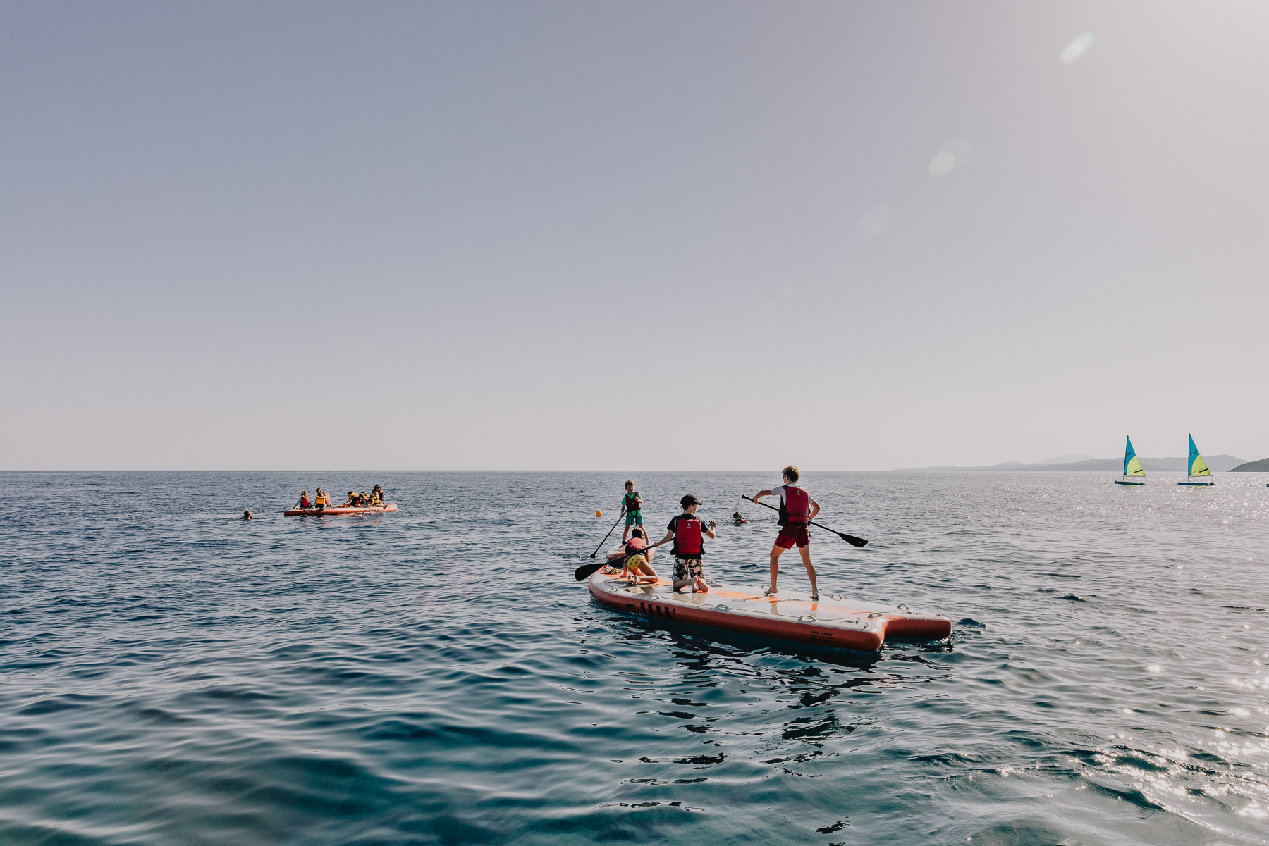 Children in red life jackets paddleboard and kayak together on open sea, with two sailing dinghies with lime-green sails visible in the distance and calm blue water stretching to the horizon.