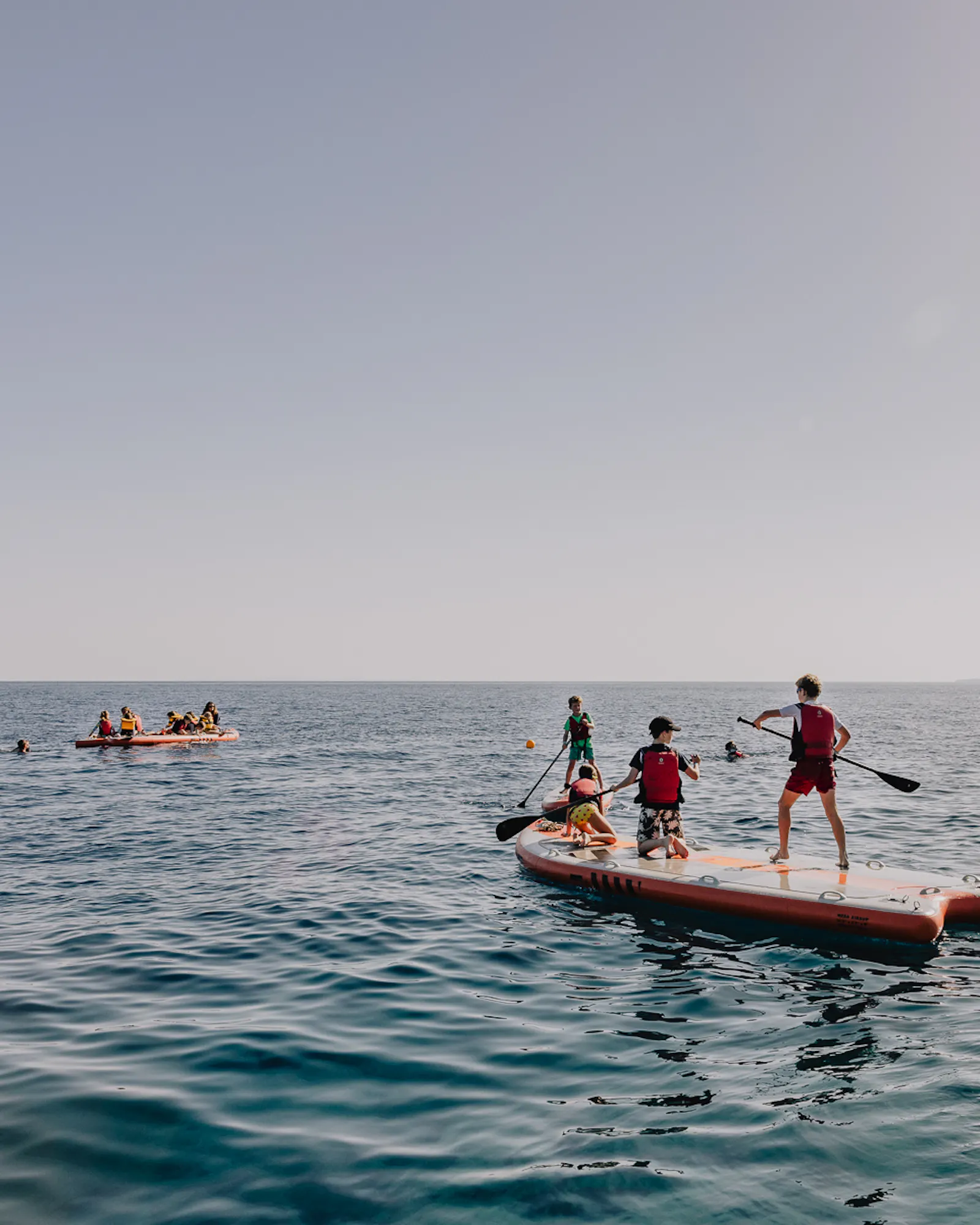 Children in red life jackets paddleboard and kayak together on open sea, with two sailing dinghies with lime-green sails visible in the distance and calm blue water stretching to the horizon.