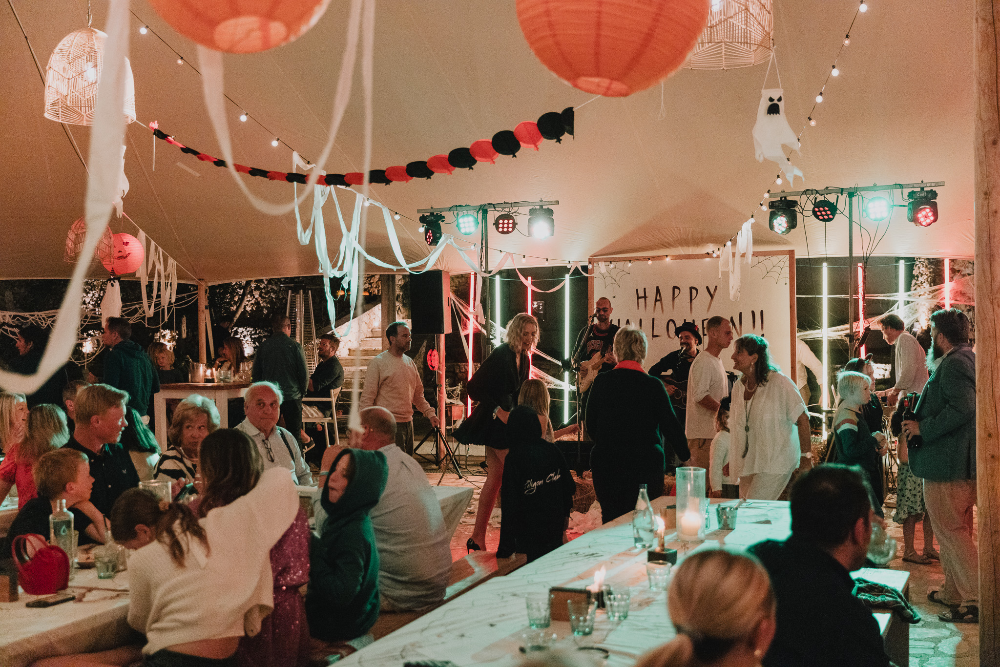 An evening scene inside a marquee decorated for Halloween with pumpkin lanterns, black-and-red bunting, and hanging ghost figures, showing a live band performing in front of a "Happy Halloween!!" sign as guests mingle and dance.