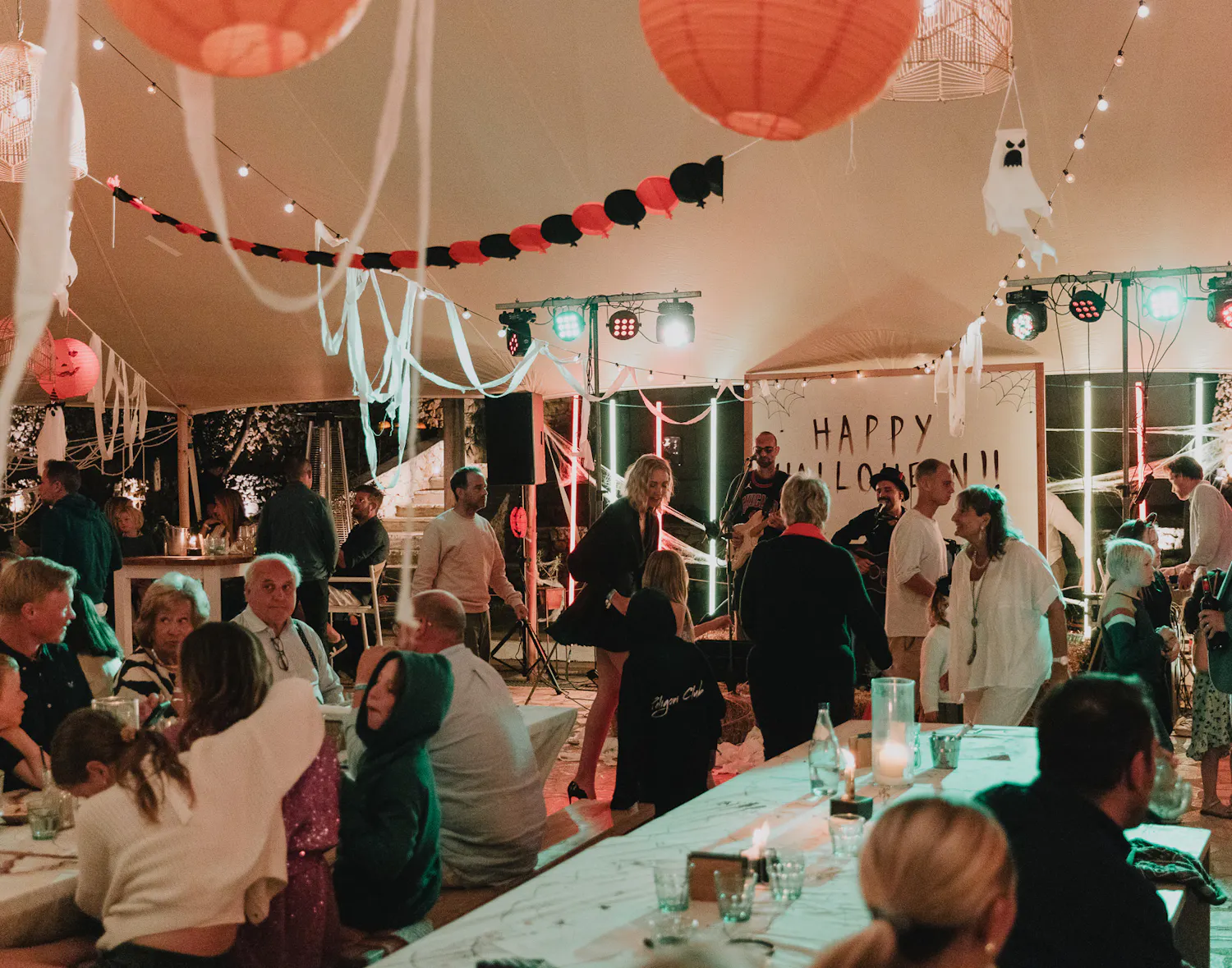 An evening scene inside a marquee decorated for Halloween with pumpkin lanterns, black-and-red bunting, and hanging ghost figures, showing a live band performing in front of a "Happy Halloween!!" sign as guests mingle and dance.