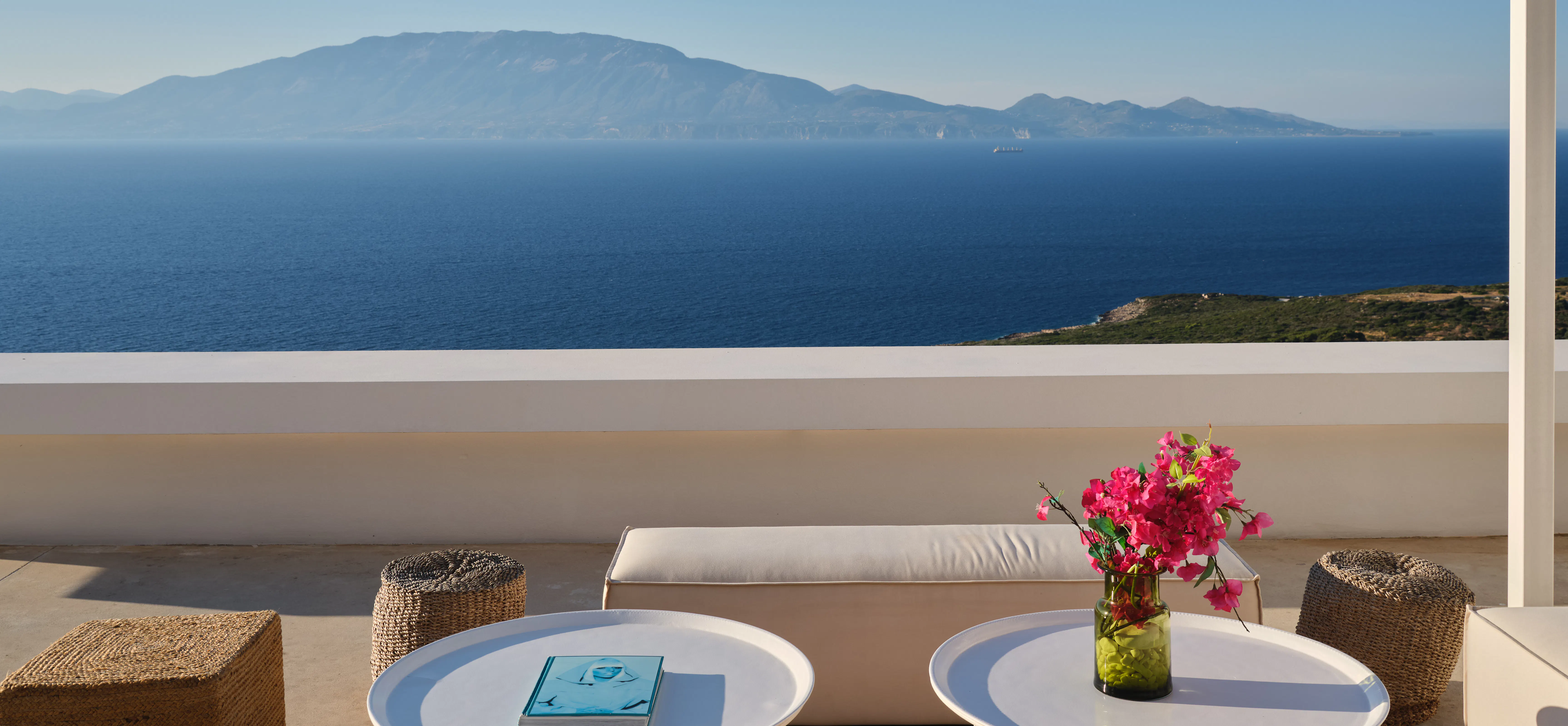 Outdoor lounge terrace with round coffee tables, a bench seat, and bougainvillea, overlooking the sea and mountains
