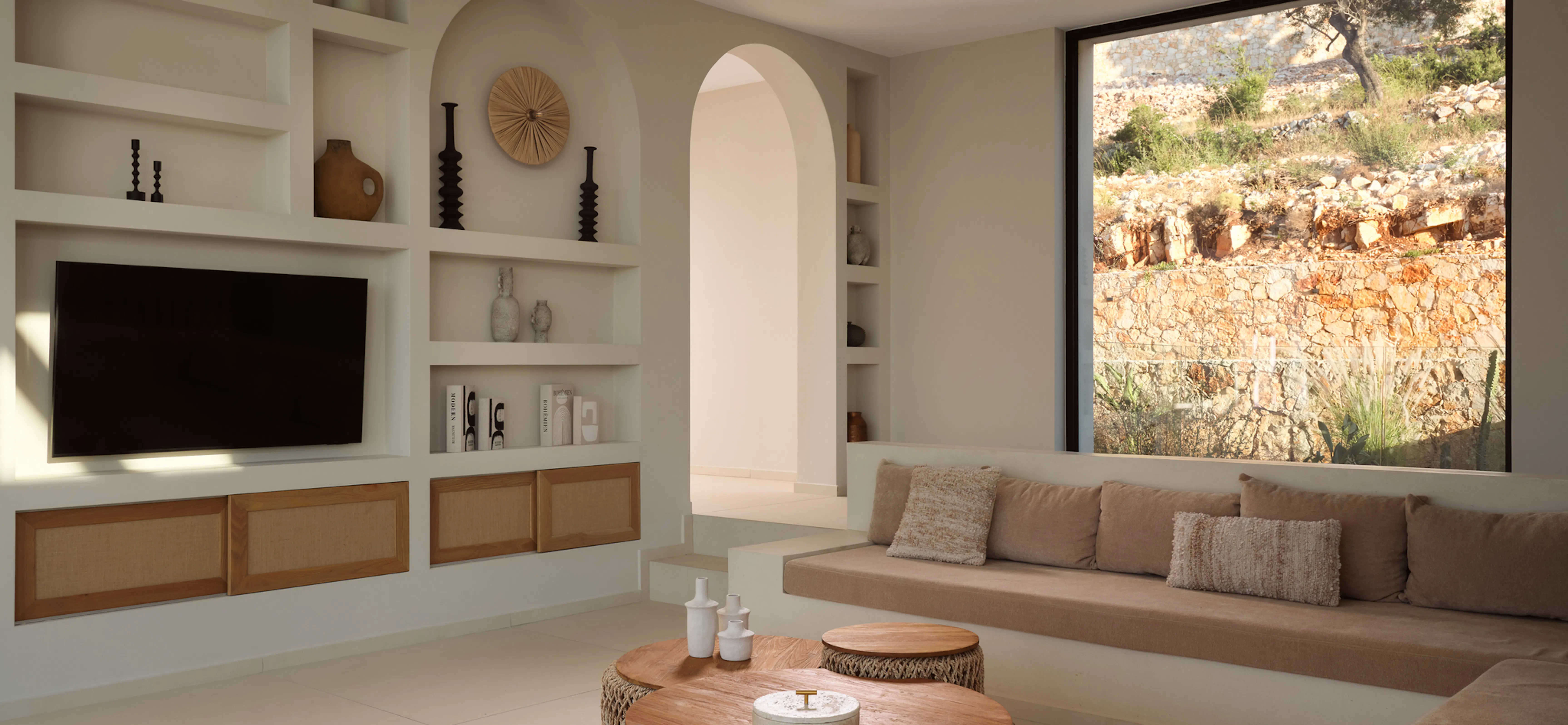 Living room with arched display shelving and mounted television, built-in banquette seating beneath a large picture window framing a rocky hillside.