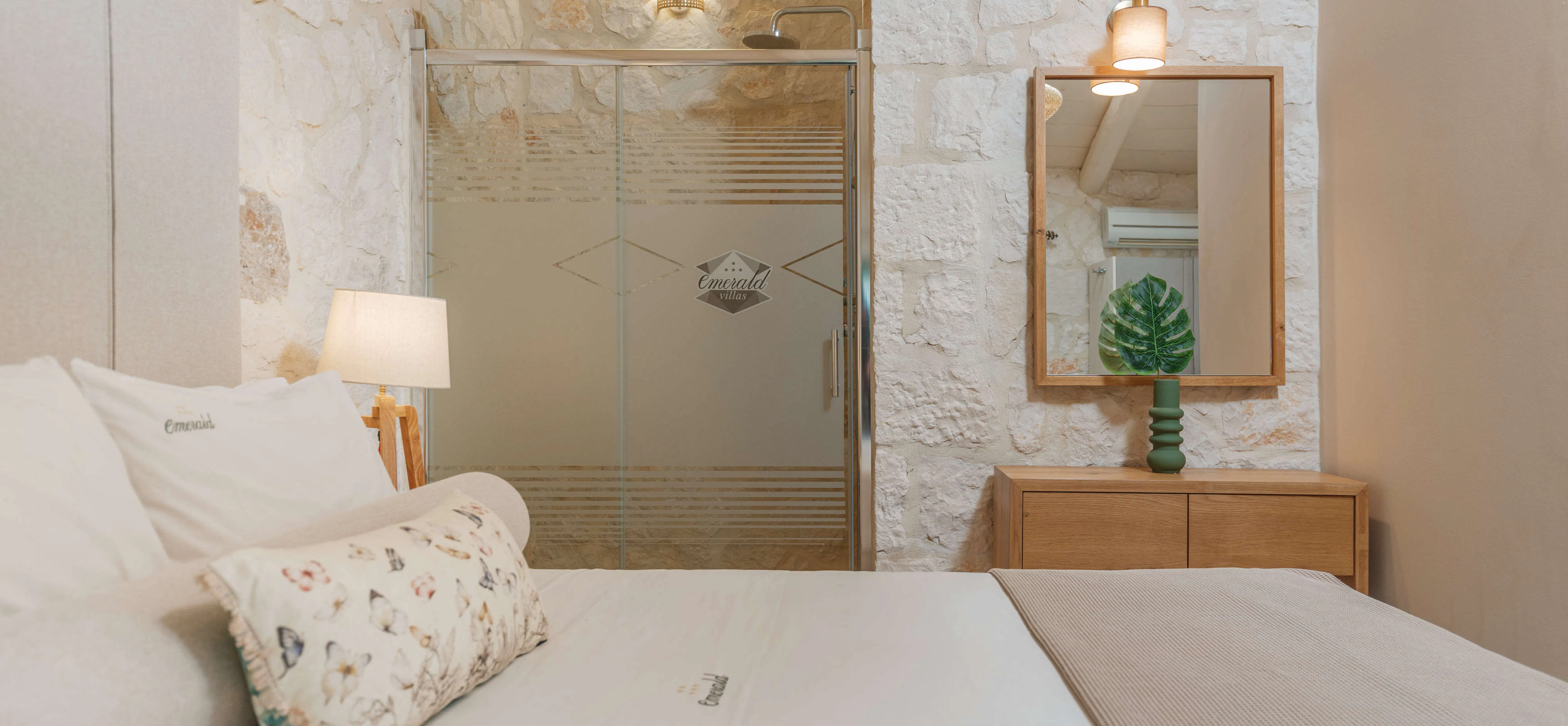 Bedroom detail showing an Emerald-branded pillow and throw against a stone wall, with a glass-enclosed shower, wooden dresser, and framed mirror reflecting a monstera leaf.