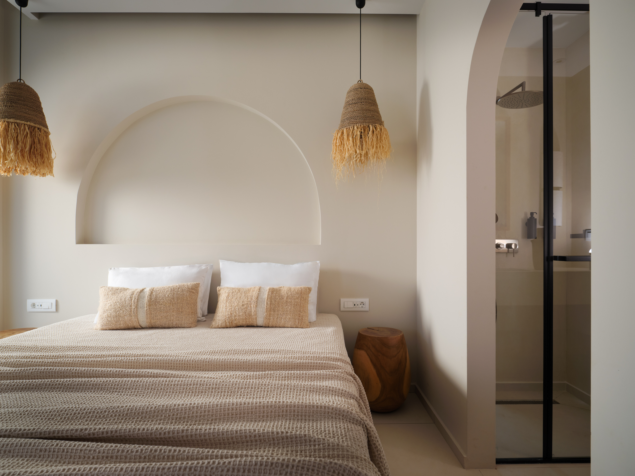 Bedroom with arched headboard niche and woven fringed pendant lights, waffle-weave linens and glimpse into an ensuite shower through a dark-framed doorway.