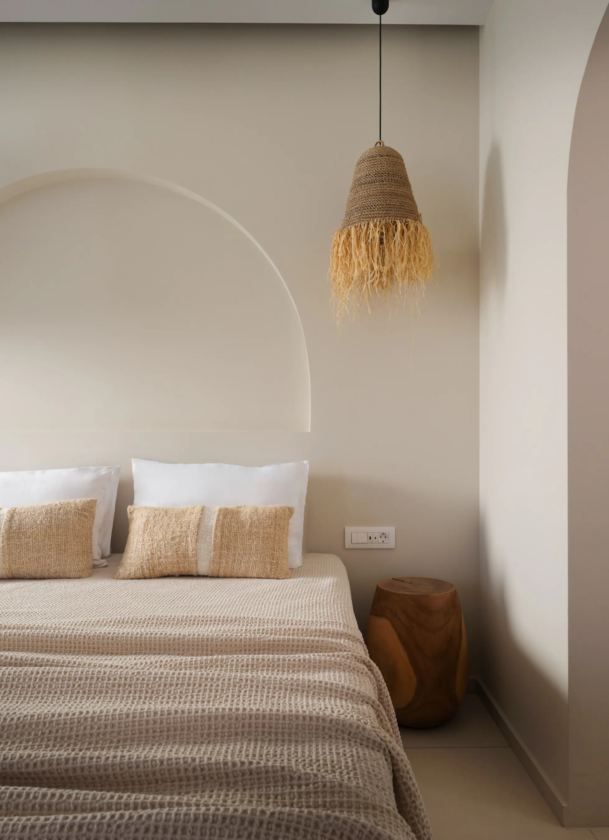 Bedroom with arched headboard niche and woven fringed pendant lights, waffle-weave linens and glimpse into an ensuite shower through a dark-framed doorway.