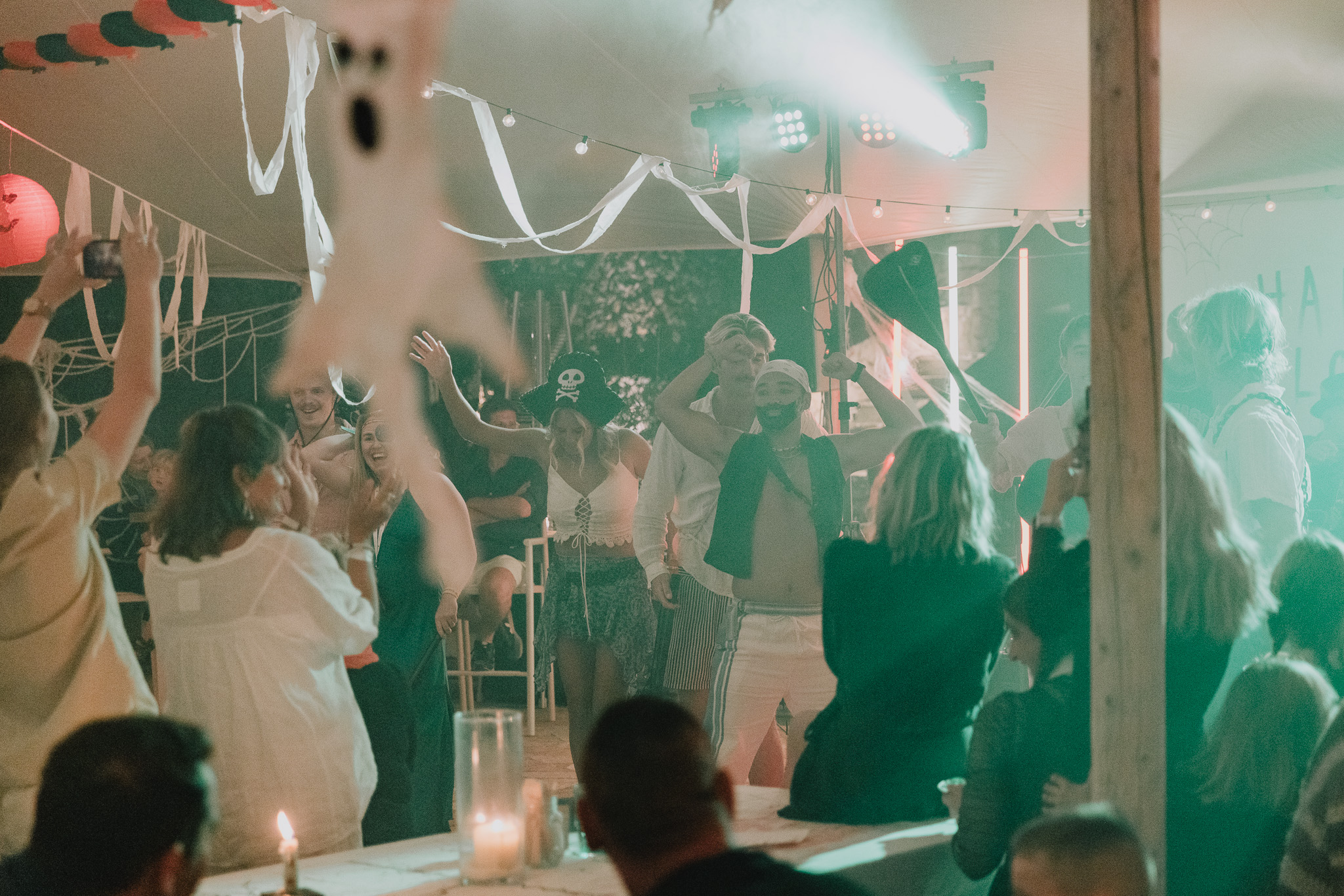 Guests dance and celebrate at a Halloween-themed evening event under a marquee decorated with white ribbon streamers, hanging pumpkin lanterns, and coloured stage lighting, with costumed partygoers including a pirate hat visible among the crowd.