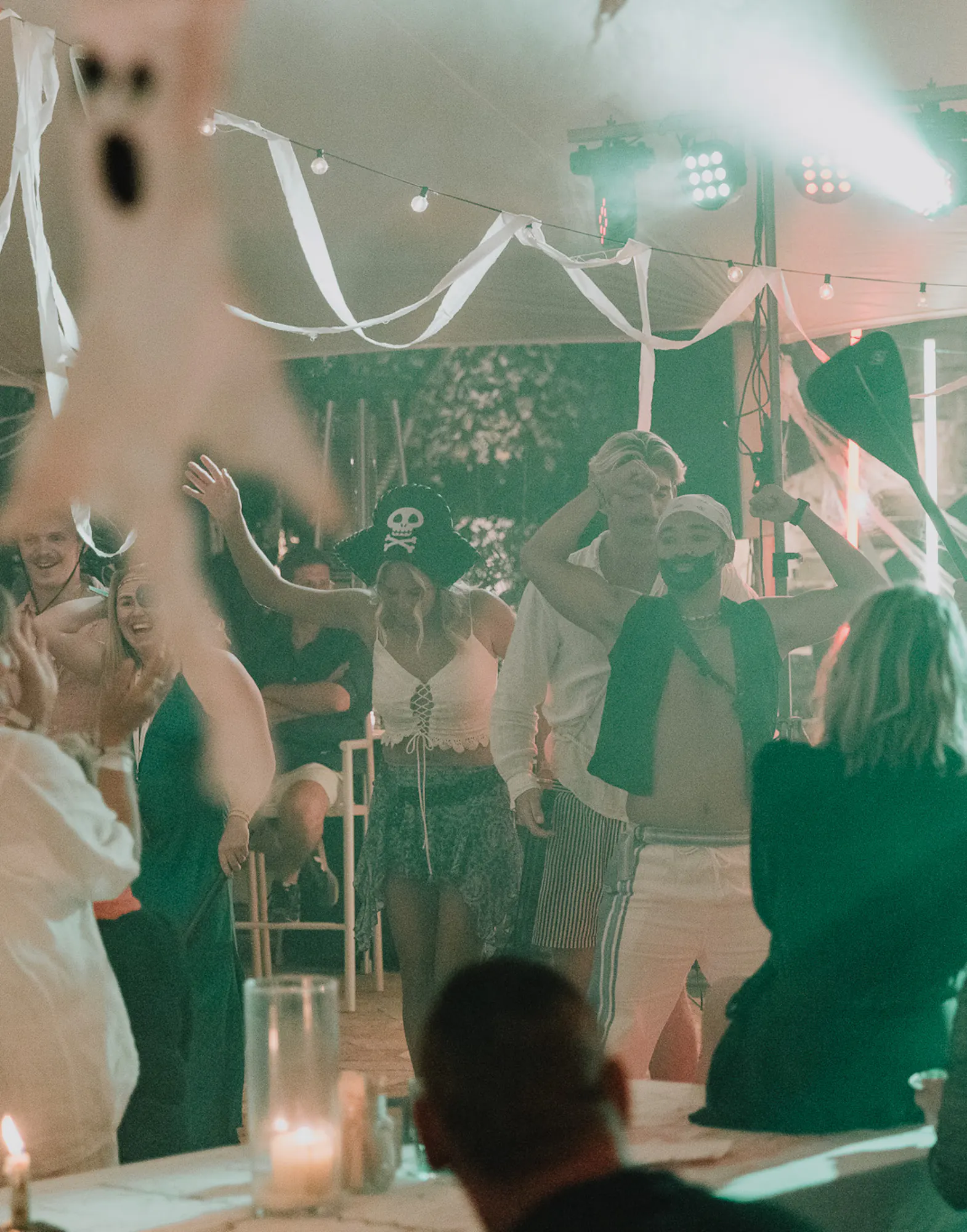 Guests dance and celebrate at a Halloween-themed evening event under a marquee decorated with white ribbon streamers, hanging pumpkin lanterns, and coloured stage lighting, with costumed partygoers including a pirate hat visible among the crowd.