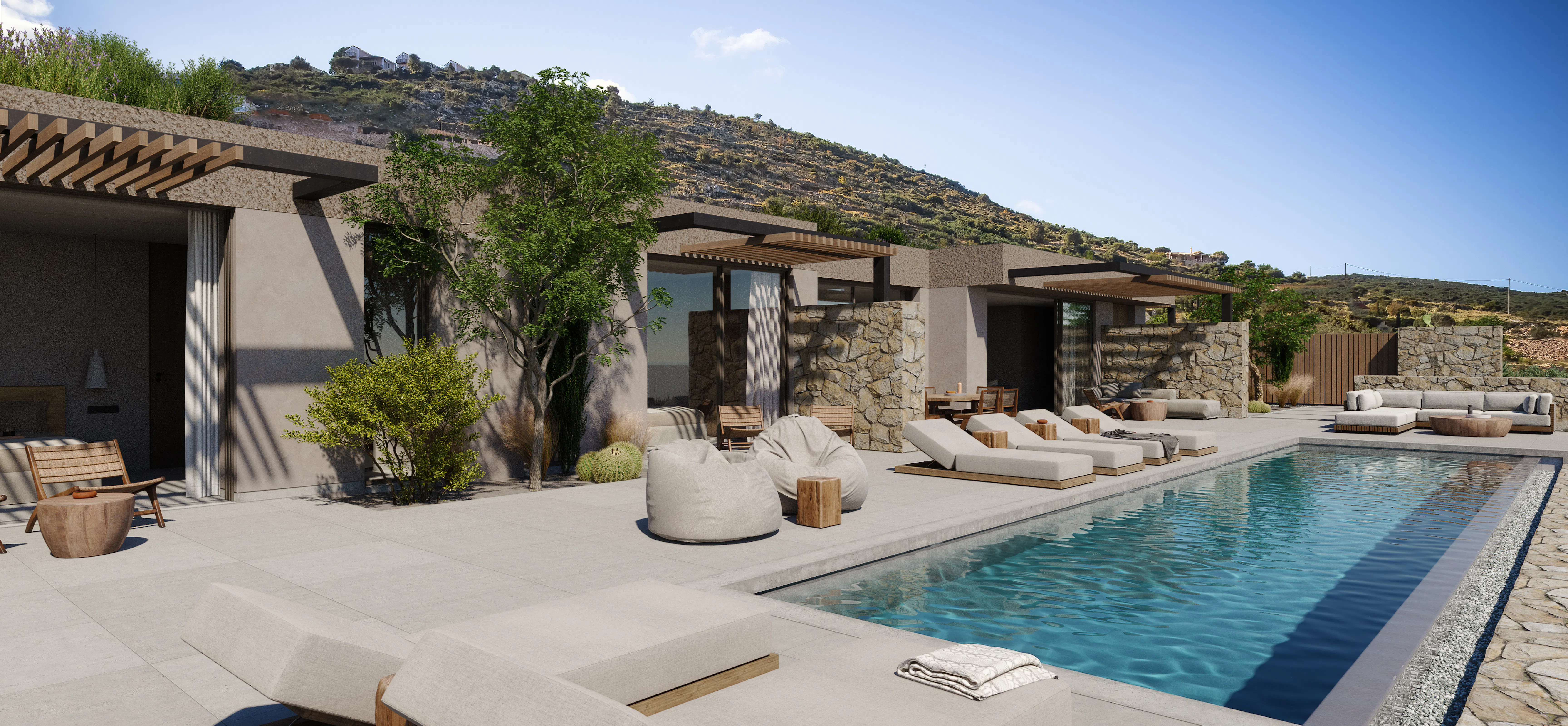 Angled view along a poolside terrace with lounge chairs, bean bags, and a stone-clad wall, backed by a scrubby hillside under a clear blue sky.