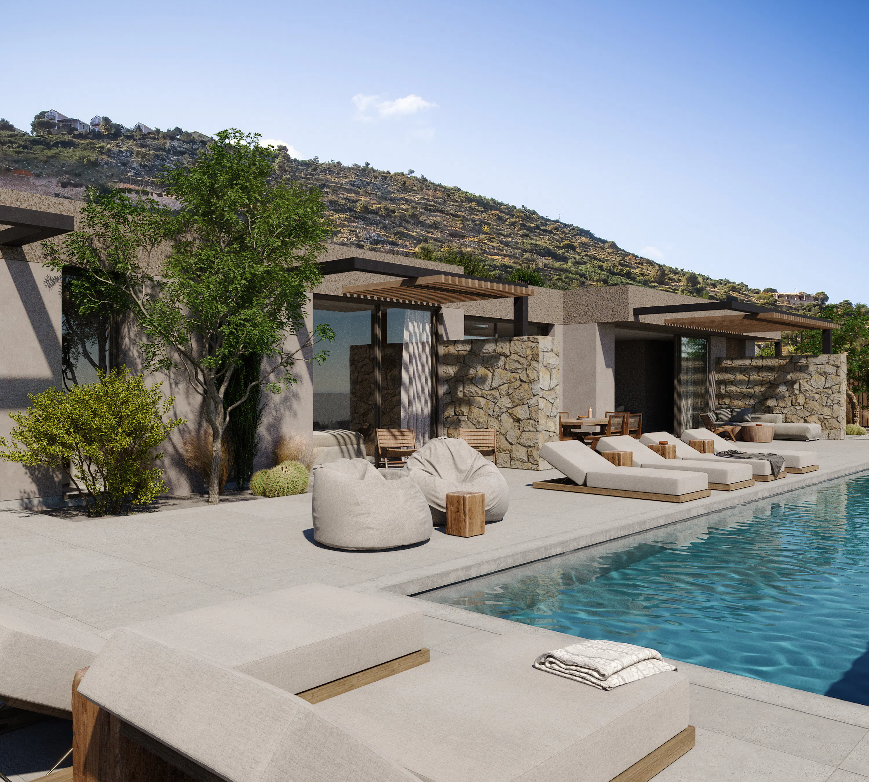 Angled view along a poolside terrace with lounge chairs, bean bags, and a stone-clad wall, backed by a scrubby hillside under a clear blue sky.