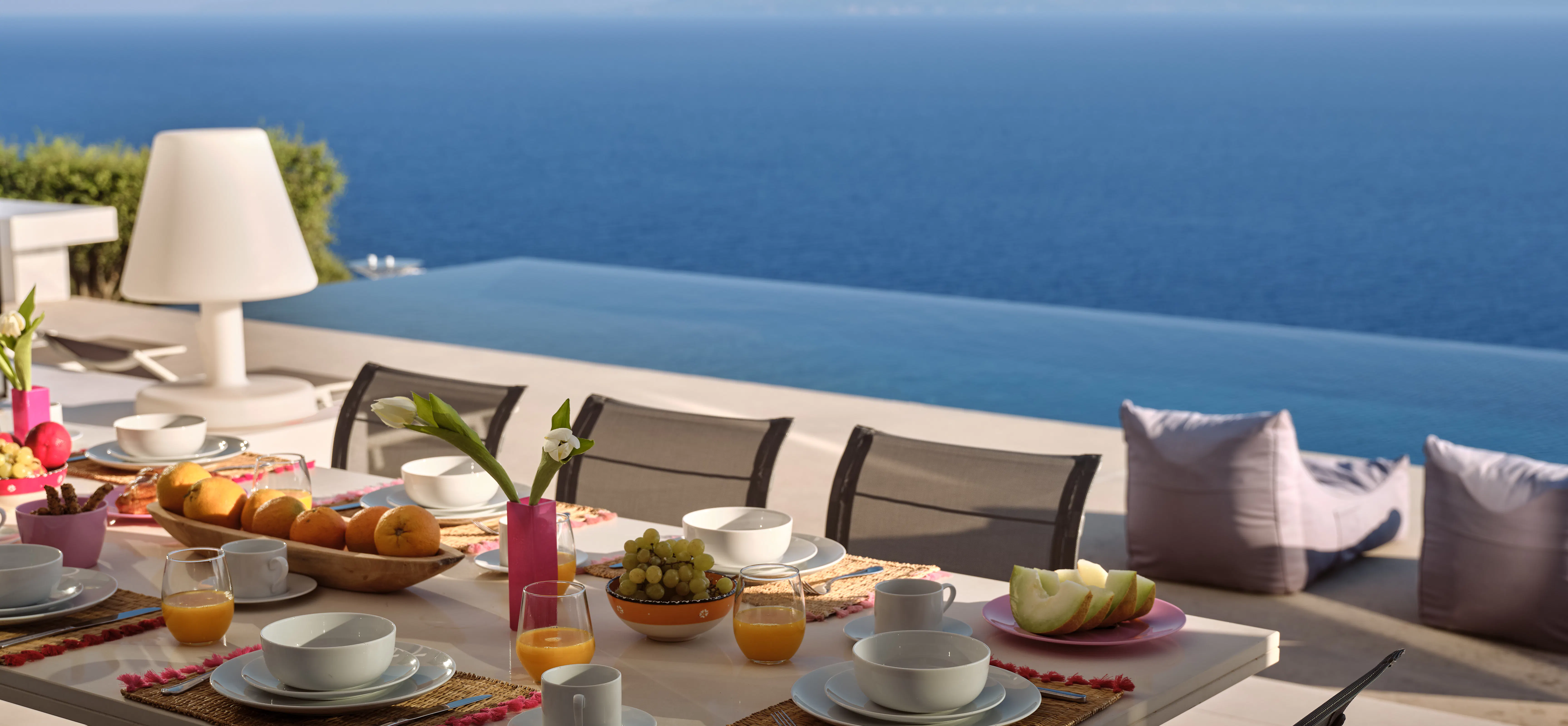Close-up of an outdoor breakfast table with pastries, fruit, orange juice, and coffee, set against a sea view