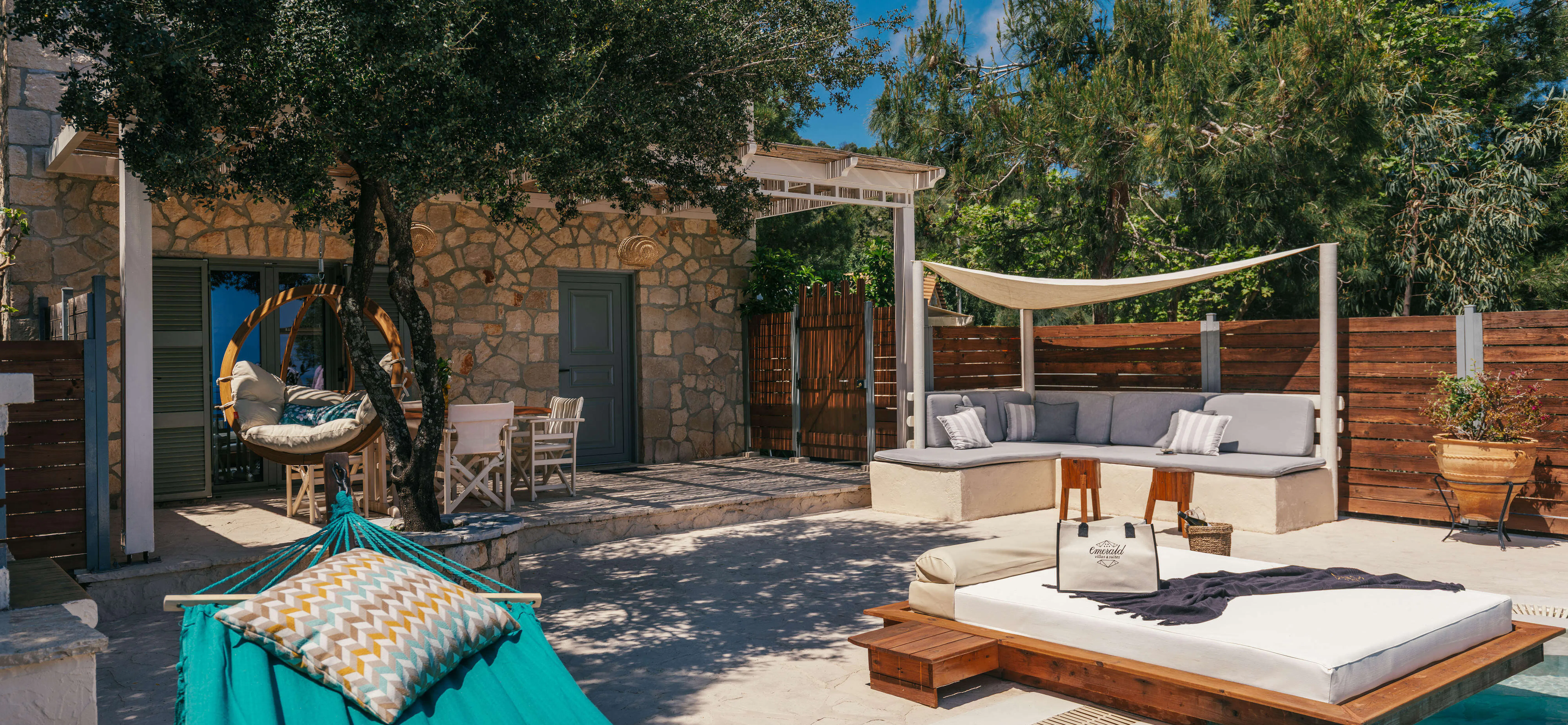 Sun-dappled poolside lounge area with a turquoise hammock, cushioned corner seating beneath a canvas sail shade, and a suspended wooden daybed extending over the pool.