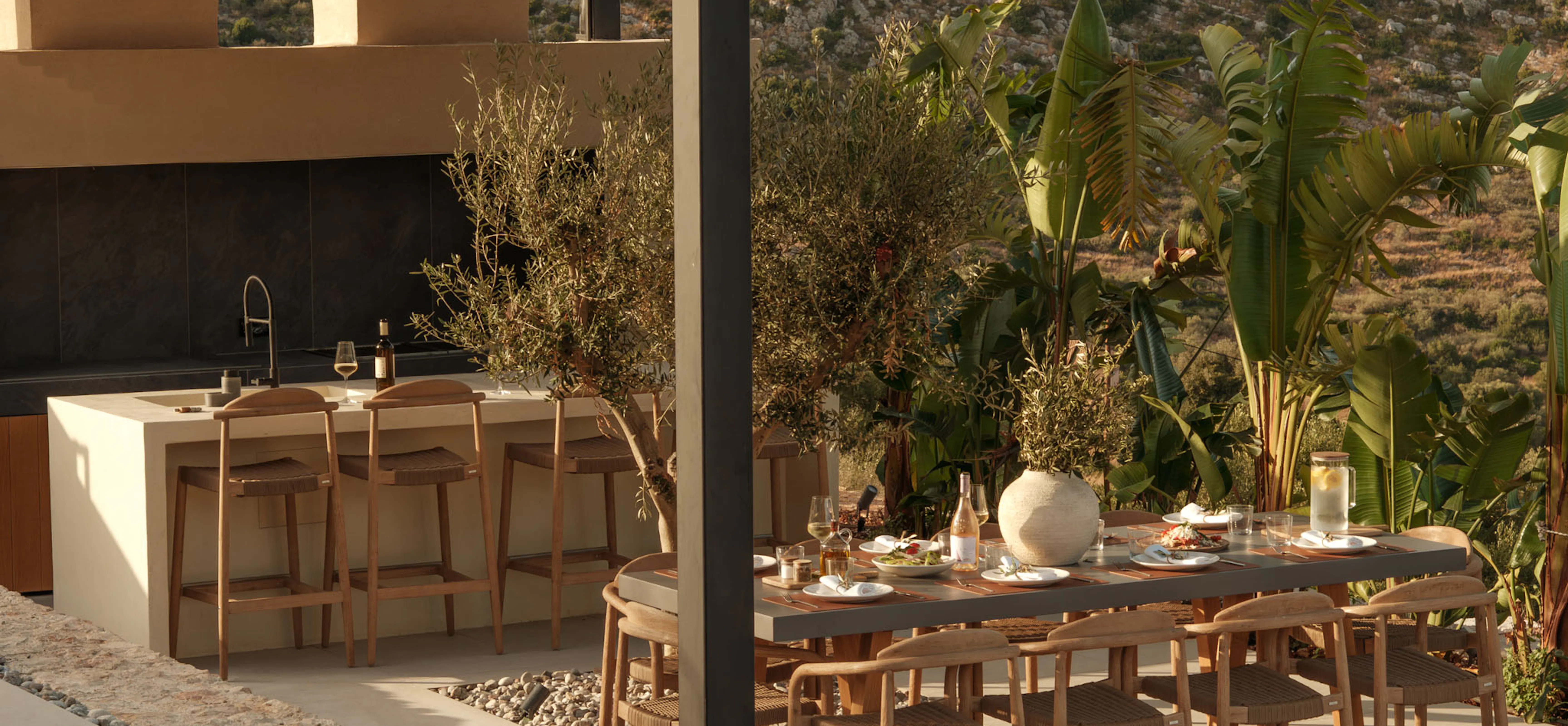Outdoor dining and kitchen pergola at The Upper House with a bar counter, wine and lemon water, and a long teak table set for lunch beneath an olive tree