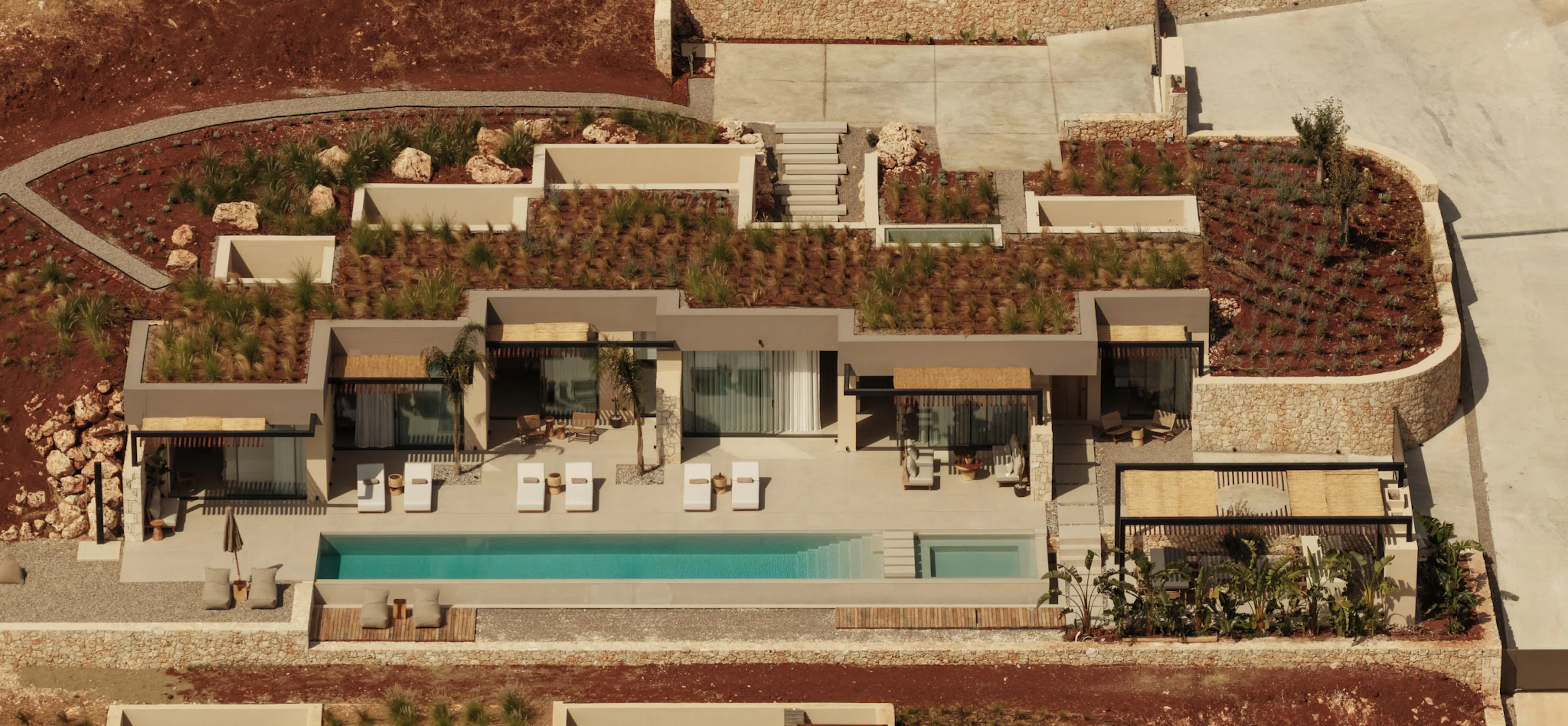 Exterior view along a pool terrace with a stone wall, loungers, and an outdoor dining area, backed by hillside.