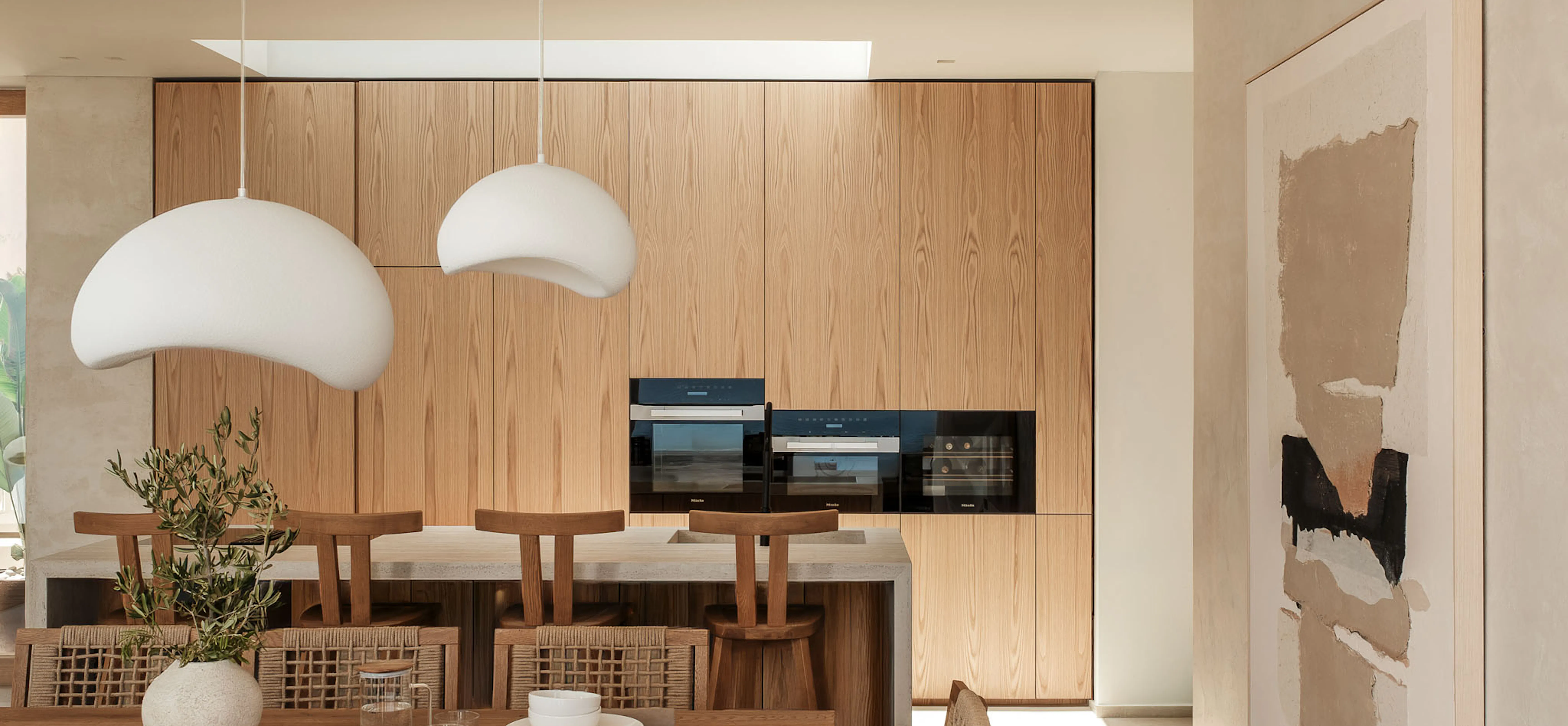 Open kitchen and dining area with two cream cocoon-shaped pendant lights over a wood table, oak cabinetry with integrated ovens, and an abstract painting on the wall.