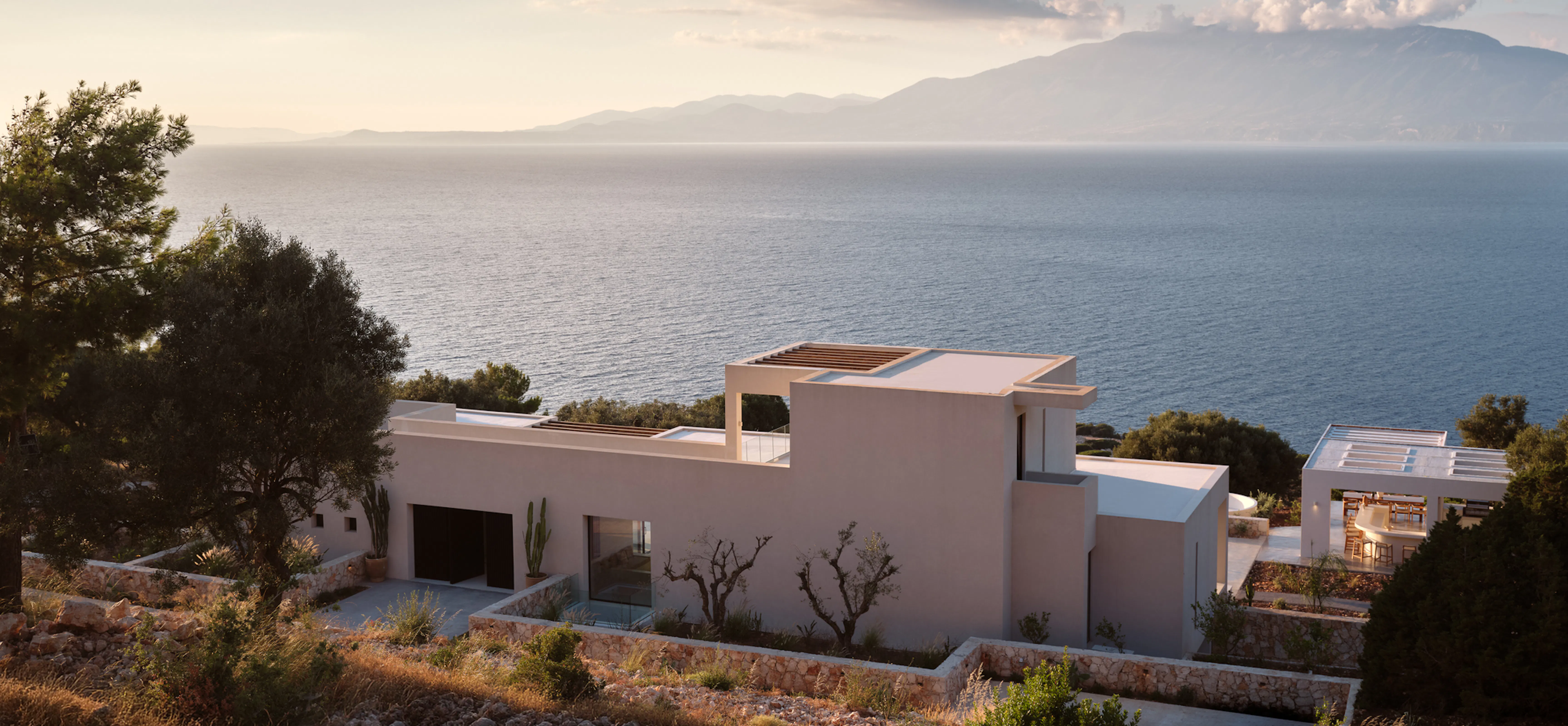 Villa exterior at golden hour, pergola-topped rooftop terrace and outdoor bar overlooking the sea beneath a hazy mountain backdrop.