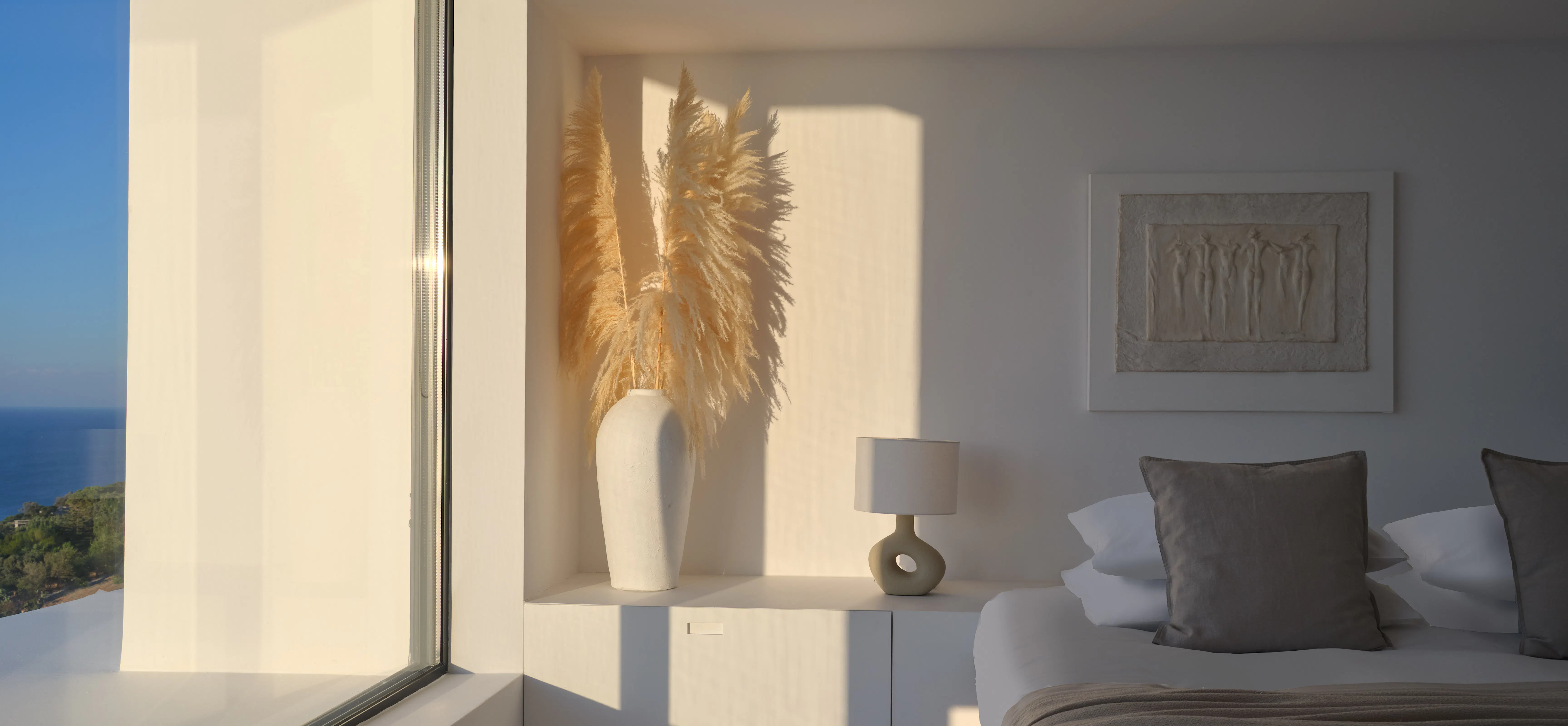 Bedroom corner with a vase of pampas grass, a round lamp, and a window with a sea view