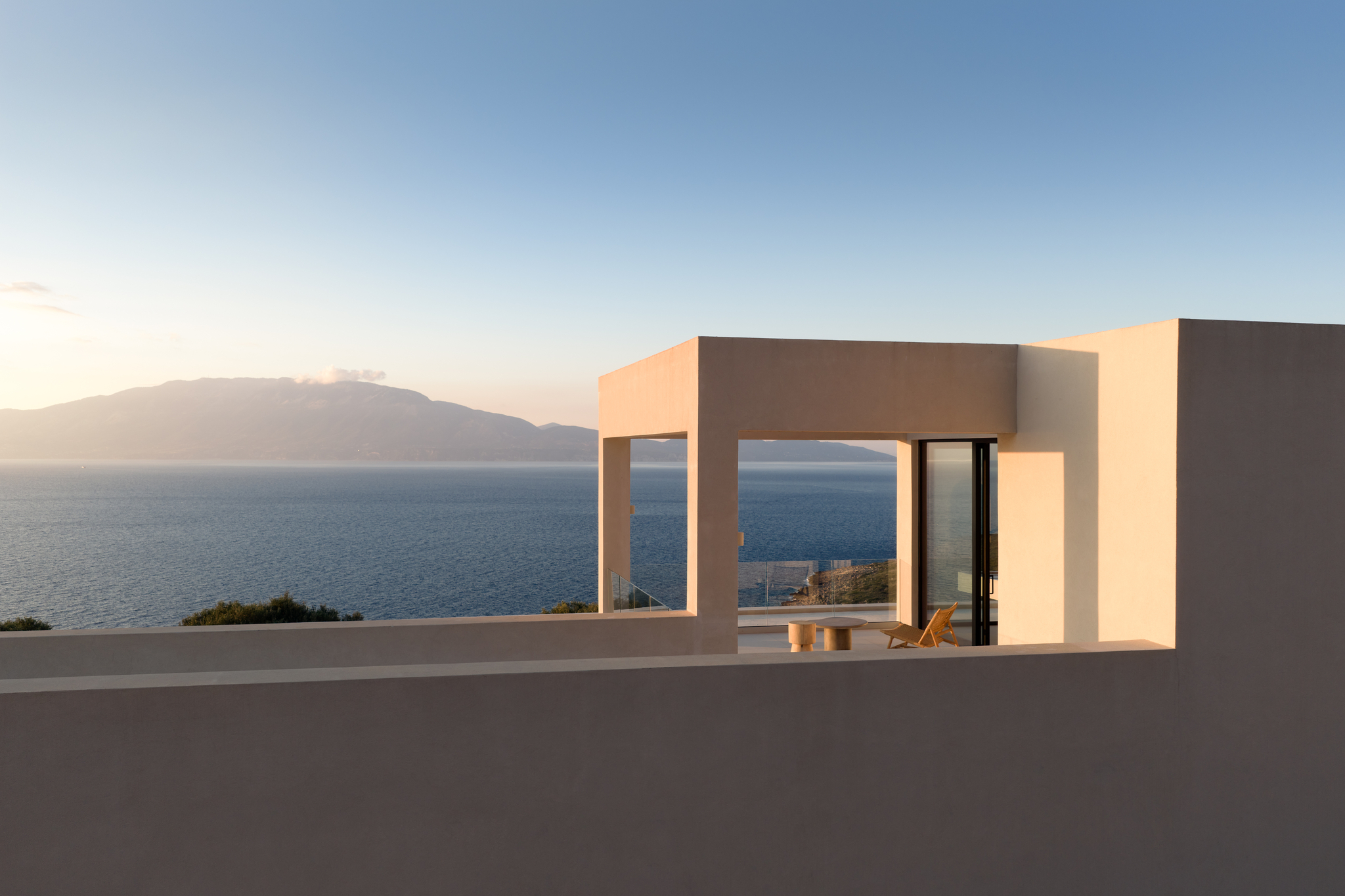 Rooftop terrace pergola at golden hour, wicker lounge chair and stump table framed by the structure's clean architectural lines, sea and mountains beyond.