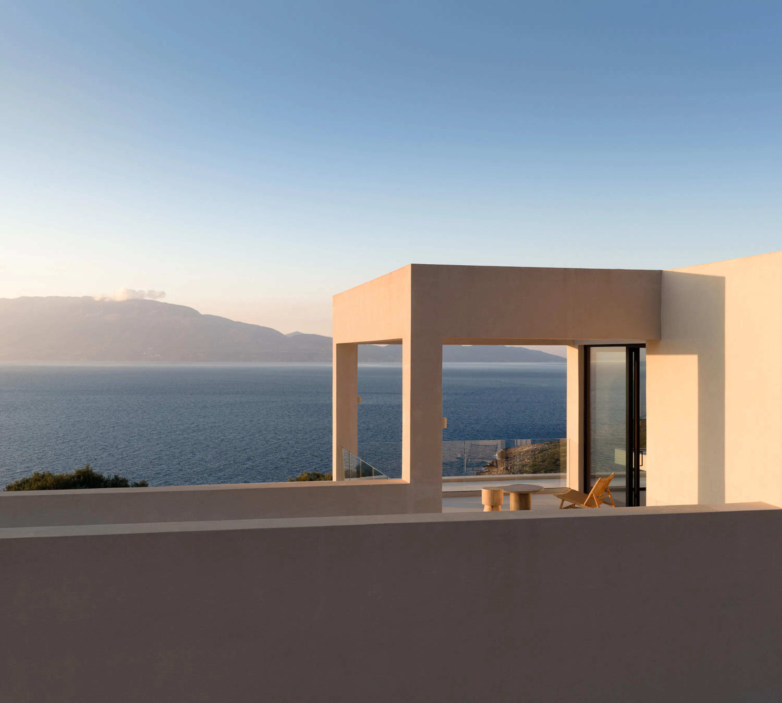 Rooftop terrace pergola at golden hour, wicker lounge chair and stump table framed by the structure's clean architectural lines, sea and mountains beyond.