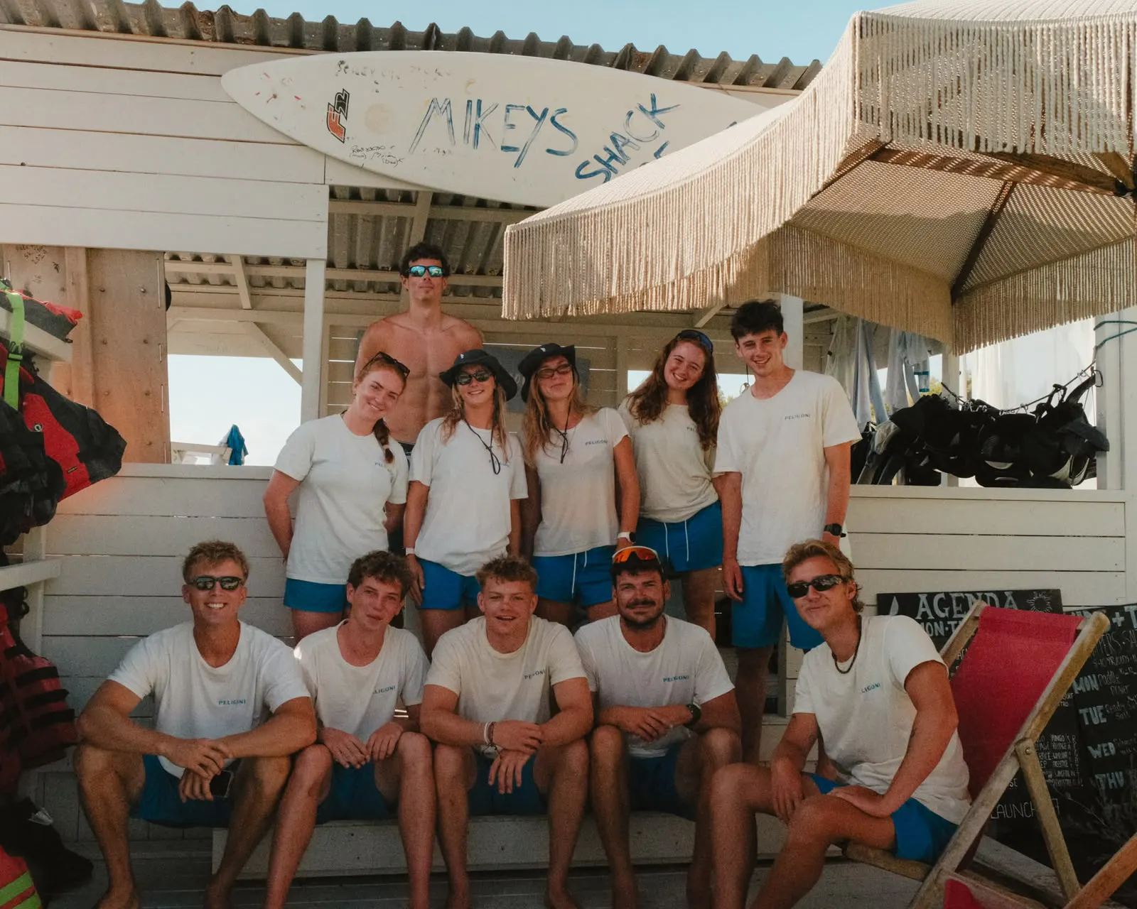 Large group of Peligoni watersports staff in white Peligoni t-shirts and blue shorts, seated and standing together beneath "Mikey's Shack" surfboard signage and a fringed cabana roof, flanked by lifejackets, sunglasses and an activities noticeboard.