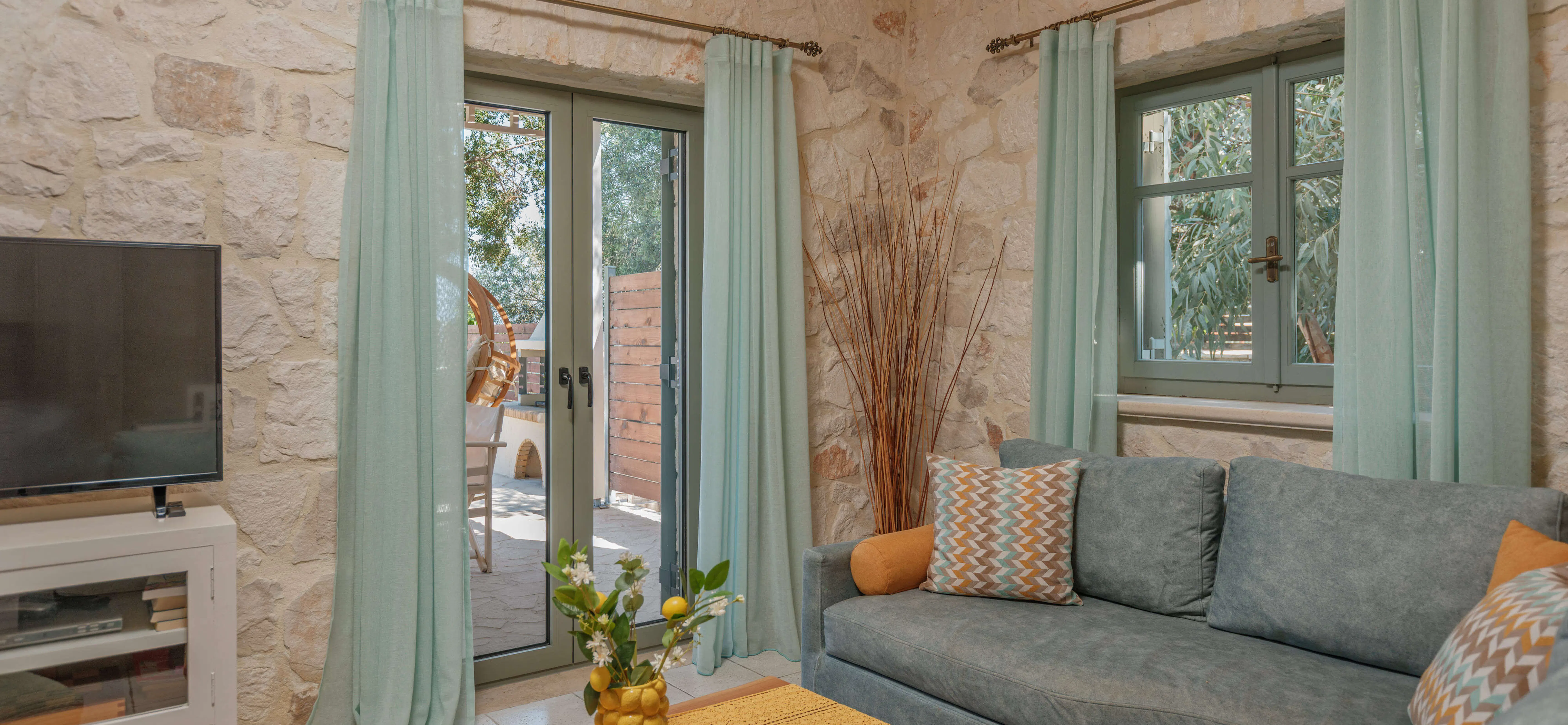 Living room corner with pale green curtains, a stone wall, and a soft blue sofa with mustard and patterned cushions arranged around a wooden coffee table.