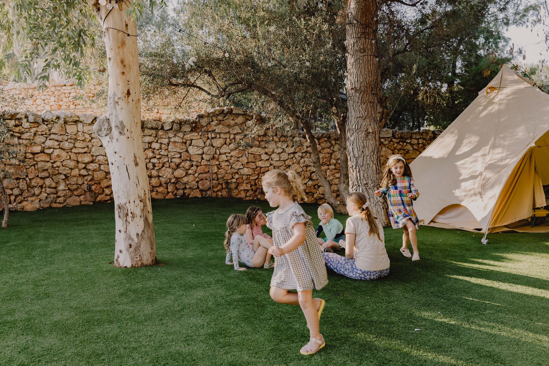 Young children in colourful summer outfits play together on artificial lawn beneath olive trees strung with fairy lights, with a canvas bell tent and a traditional dry-stone wall forming the backdrop.