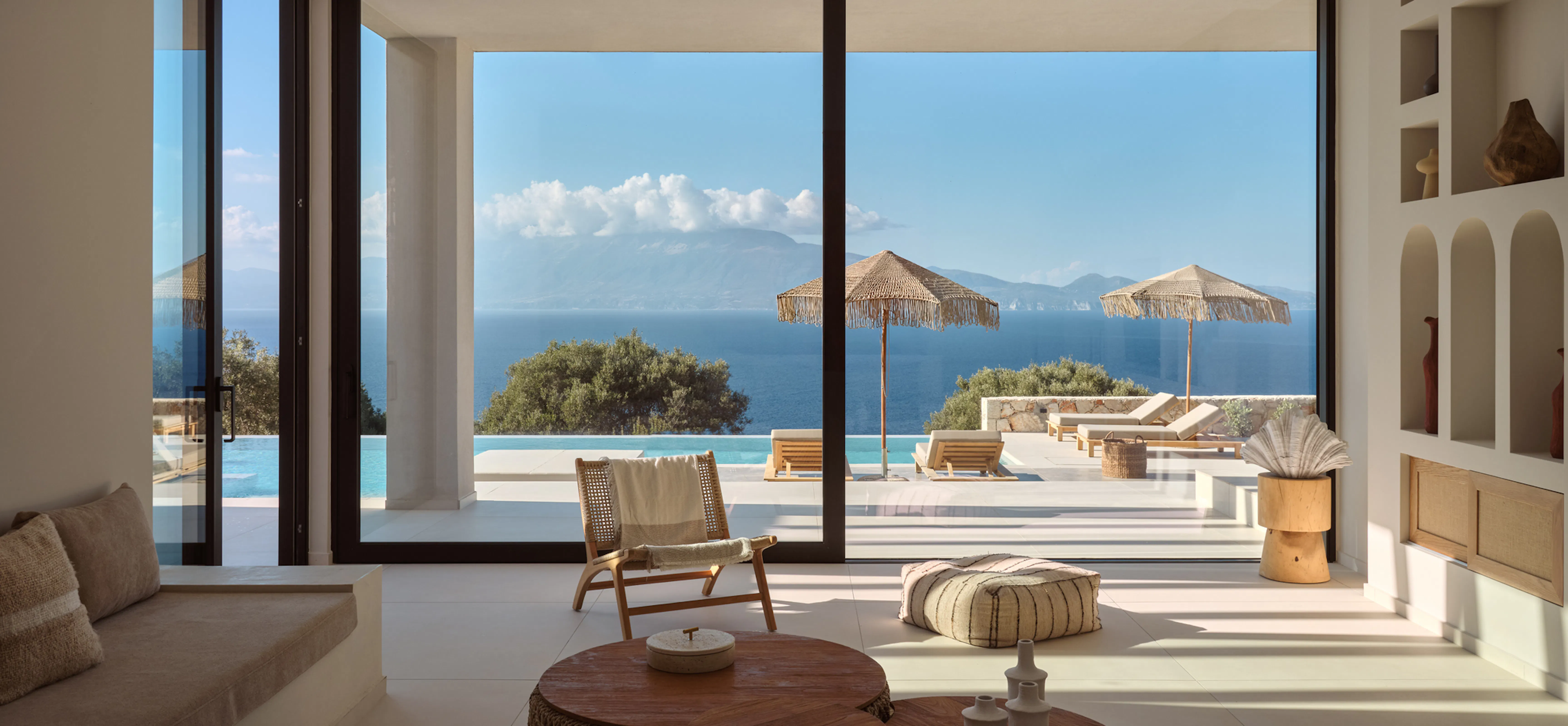 Open-plan living space with cane armchair and layered round wooden coffee tables, sliding glass doors revealing the pool terrace and fringed umbrellas beyond.