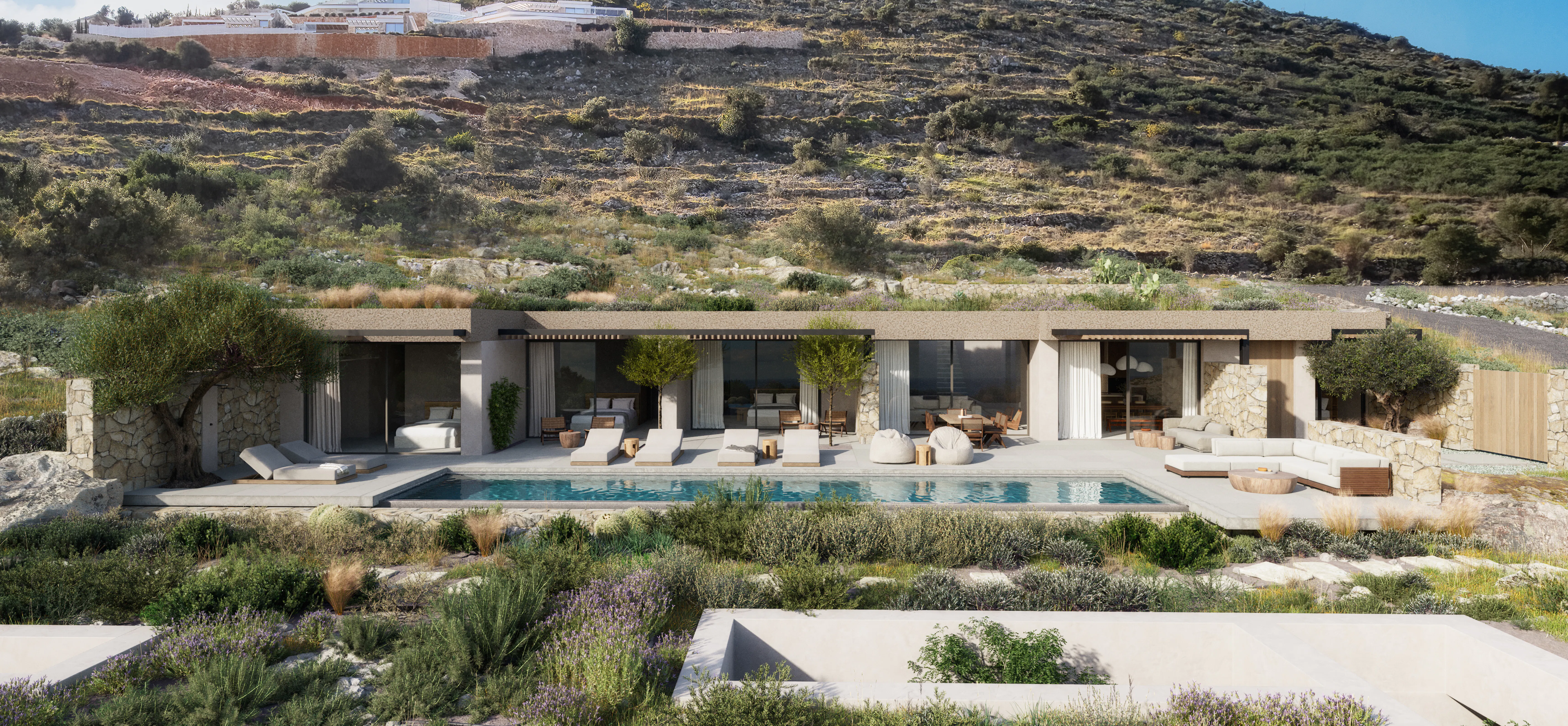 Wide exterior view of a single-story stone-and-render villa with a long lap pool, sun loungers, and outdoor seating, set beneath a terraced hillside with hilltop buildings above.