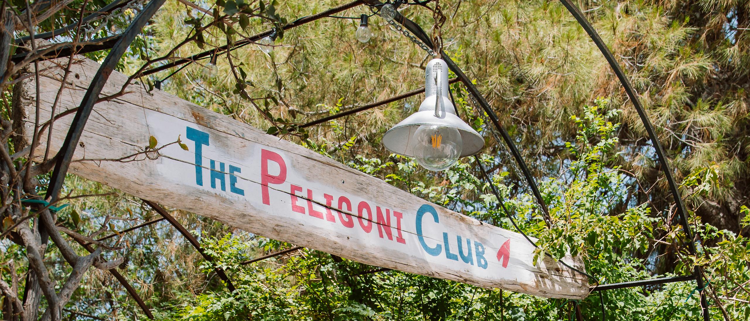An inviting entrance to The Peligoni Club featuring weathered vertical wooden planks forming a gate beneath a metal archway. A hand-painted sign reading 'The Peligoni Club' in turquoise and pink lettering hangs from the arch. The entrance is framed by lush green trees and foliage, with dappled sunlight creating shadows on the stone pathway leading through the gate. The rustic, natural aesthetic suggests a Mediterranean or coastal garden setting.