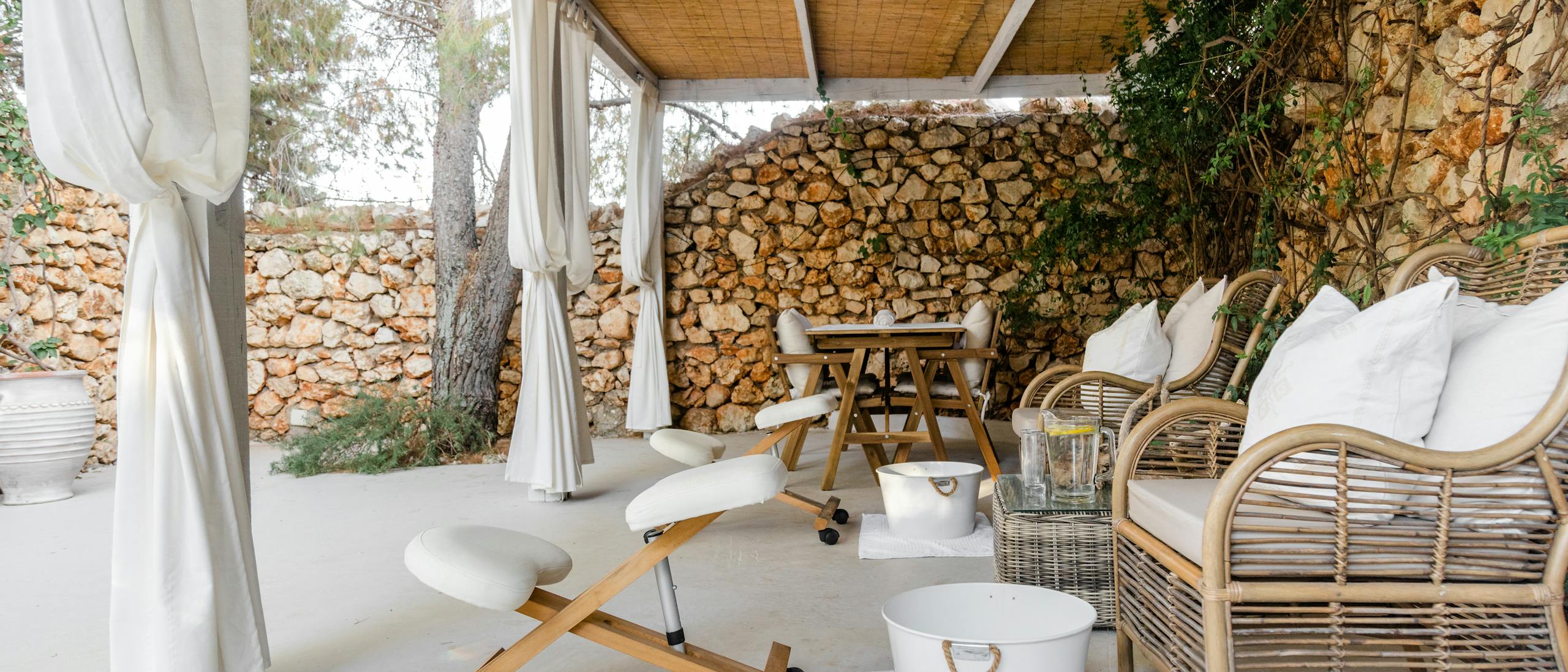 A serene outdoor spa relaxation area featuring a covered terrace with natural bamboo reed ceiling and white wooden beams. The space includes a white cushioned massage chair with wooden frame on wheels, rattan lounge chairs with plush white pillows, and a white foot bath basin with rope handles. The backdrop showcases rustic stone walls with climbing vines and Mediterranean vegetation. A small wooden side table sits between seating areas, creating an intimate wellness retreat atmosphere.