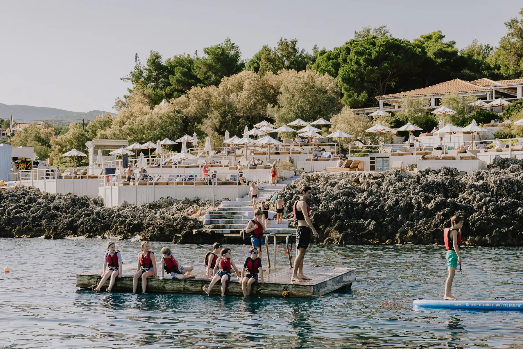 Children in red life jackets sit along a floating wooden pontoon in a rocky cove while an instructor stands ready to assist, with a beach club's white umbrellas, loungers, and stone steps visible on the shoreline behind them.