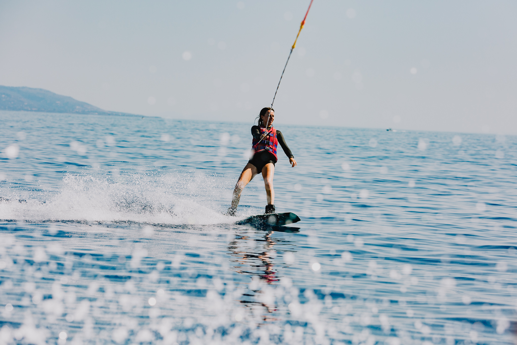 A person wakeboarding across calm turquoise water, creating a spray of white water behind them. They wear a black wetsuit with pink accents and are being pulled by a tow rope visible above. Mountains or hills are visible in the misty distance along the horizon. The water's surface reflects the overcast sky.