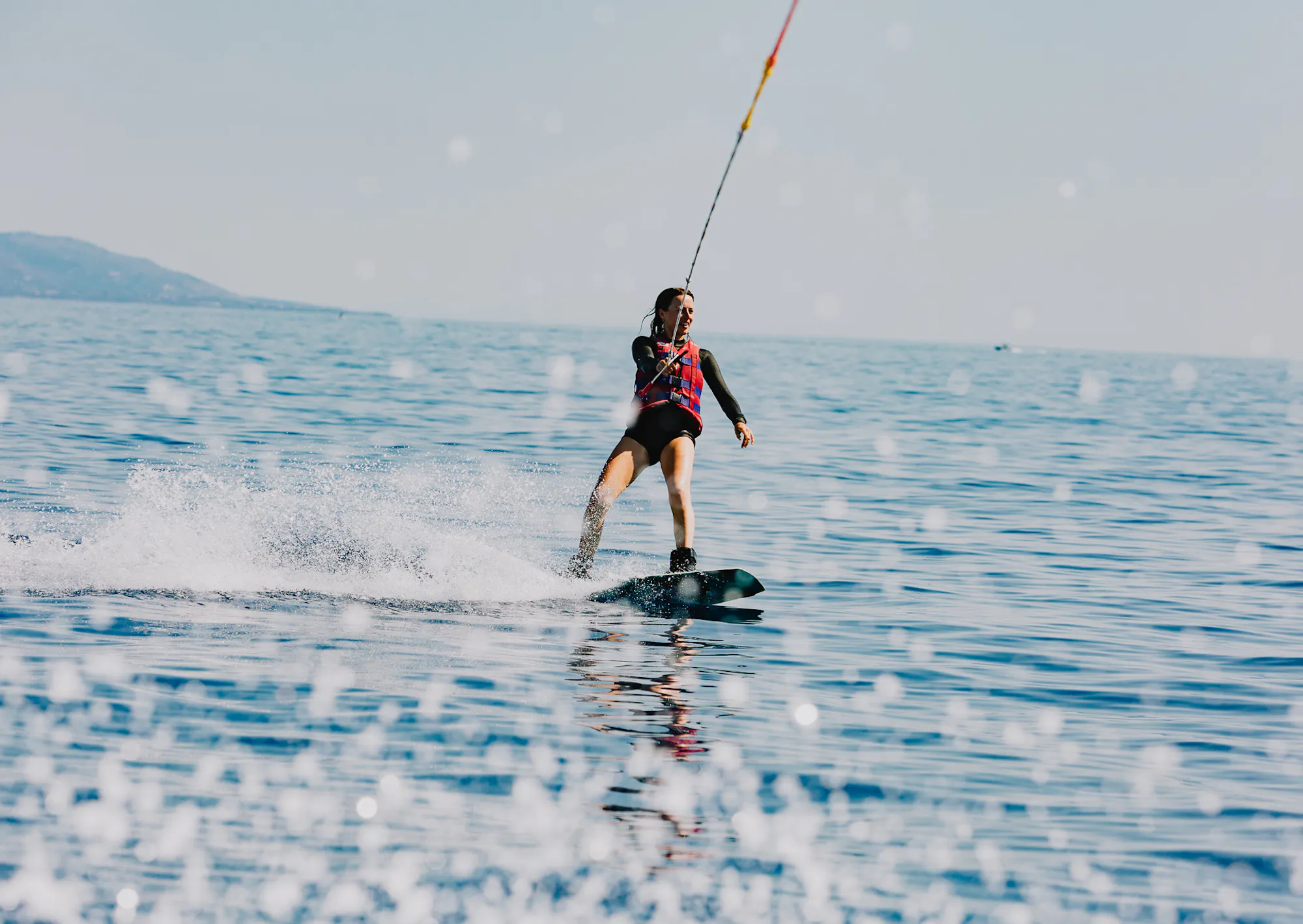 A person wakeboarding across calm turquoise water, creating a spray of white water behind them. They wear a black wetsuit with pink accents and are being pulled by a tow rope visible above. Mountains or hills are visible in the misty distance along the horizon. The water's surface reflects the overcast sky.