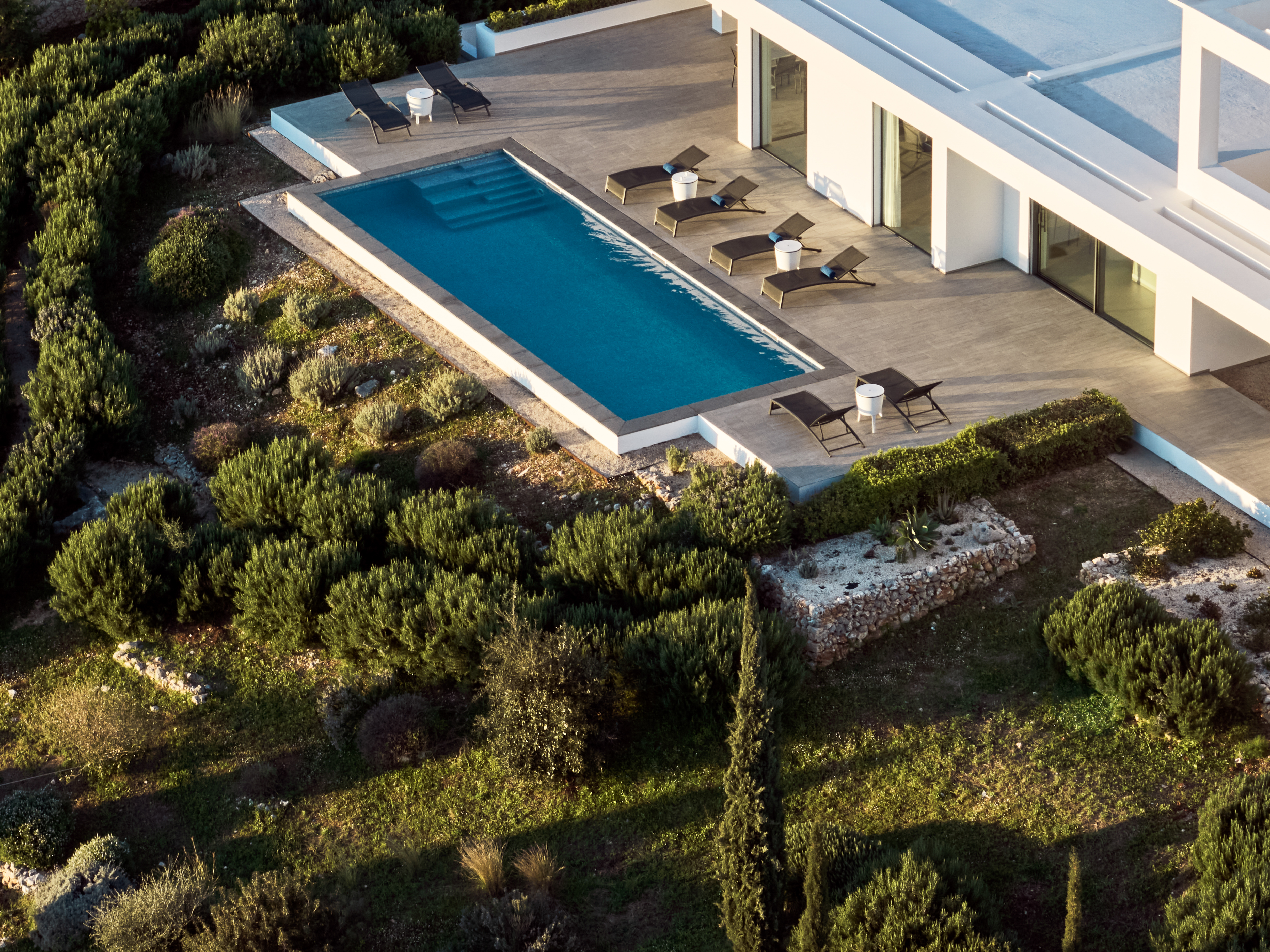 Aerial view of rectangular infinity pool with dark loungers, white side tables, and wood deck surrounded by Mediterranean scrubland.