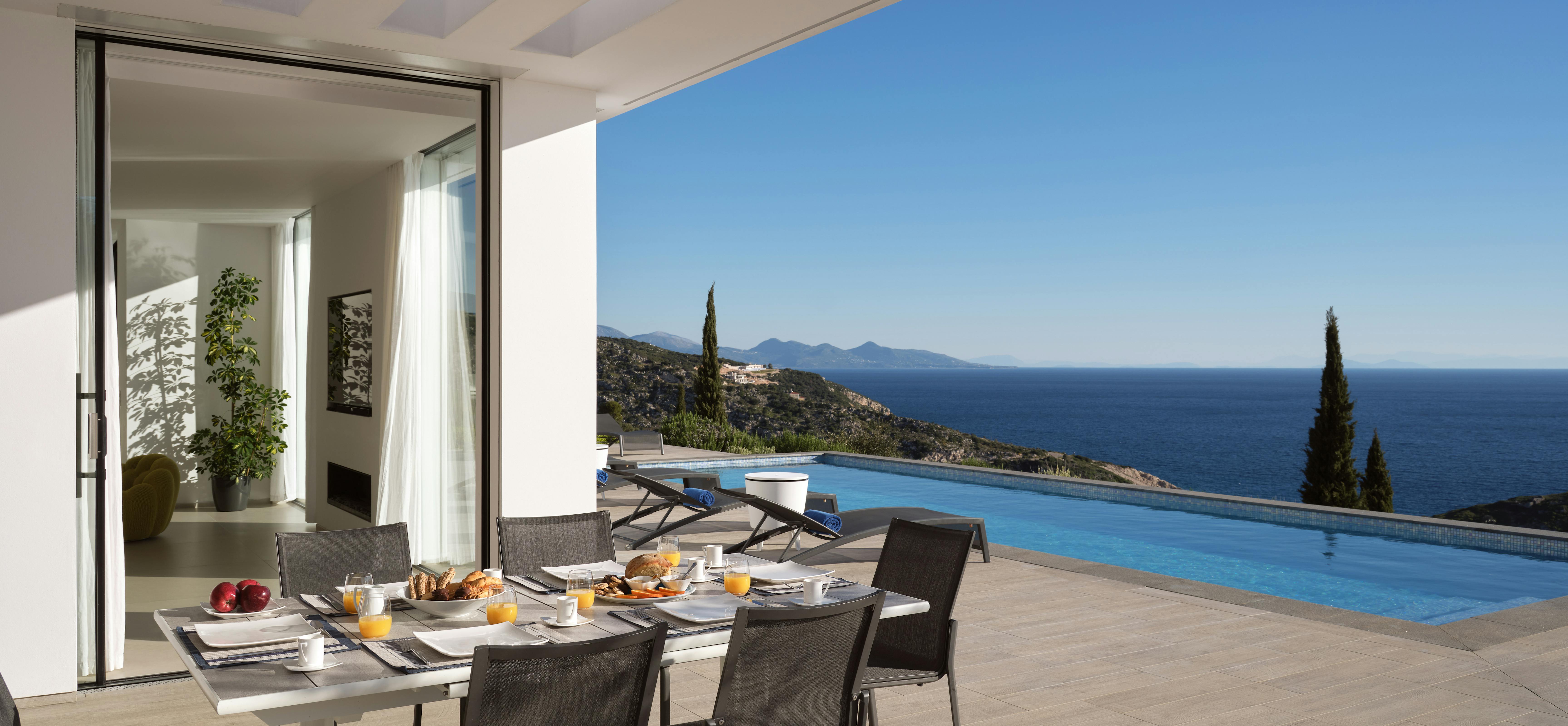 Covered breakfast terrace with modern chairs, table set with fresh fruit and pastries, infinity pool, and cypress-studded coastal panorama beyond.