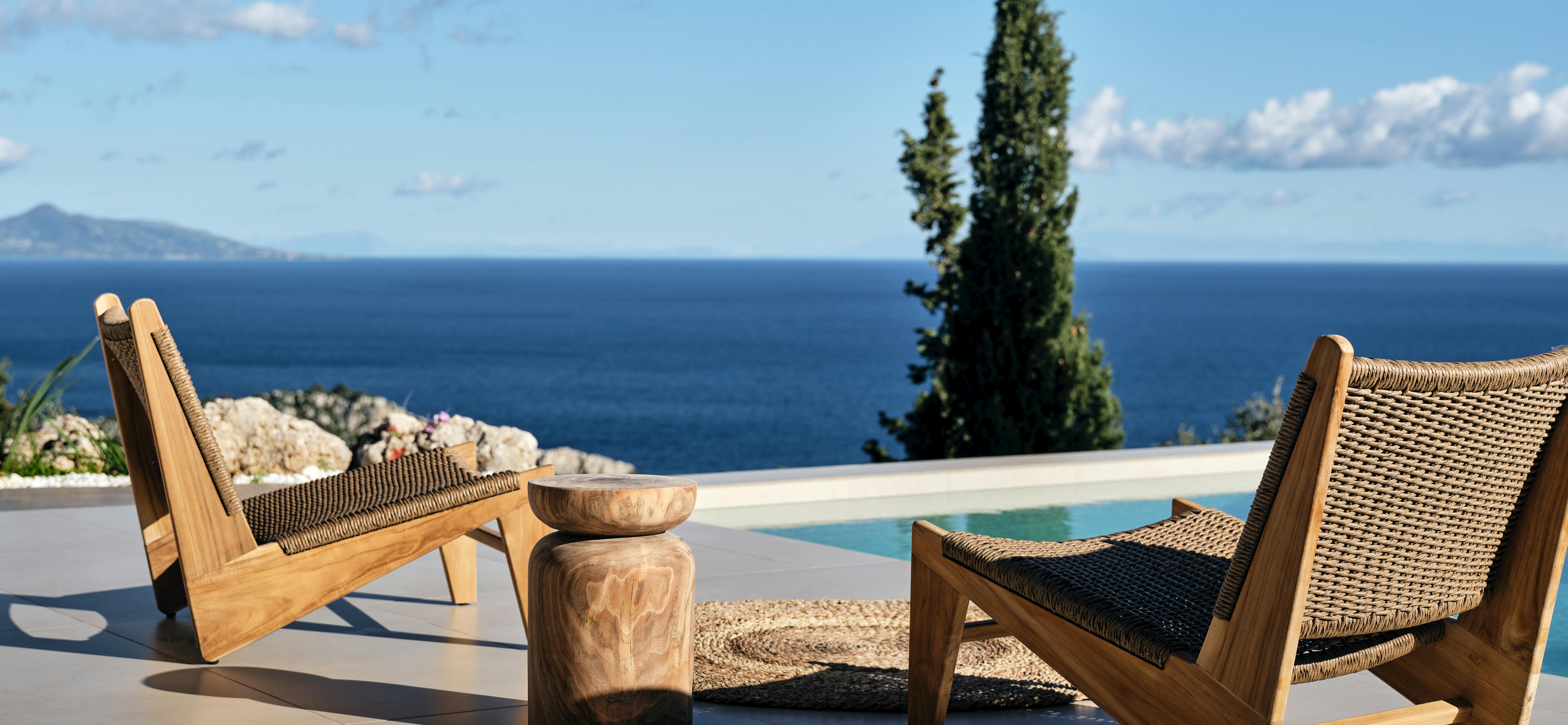 Pair of modern wood-and-rope lounge chairs with carved side table on infinity pool deck facing endless blue sea.