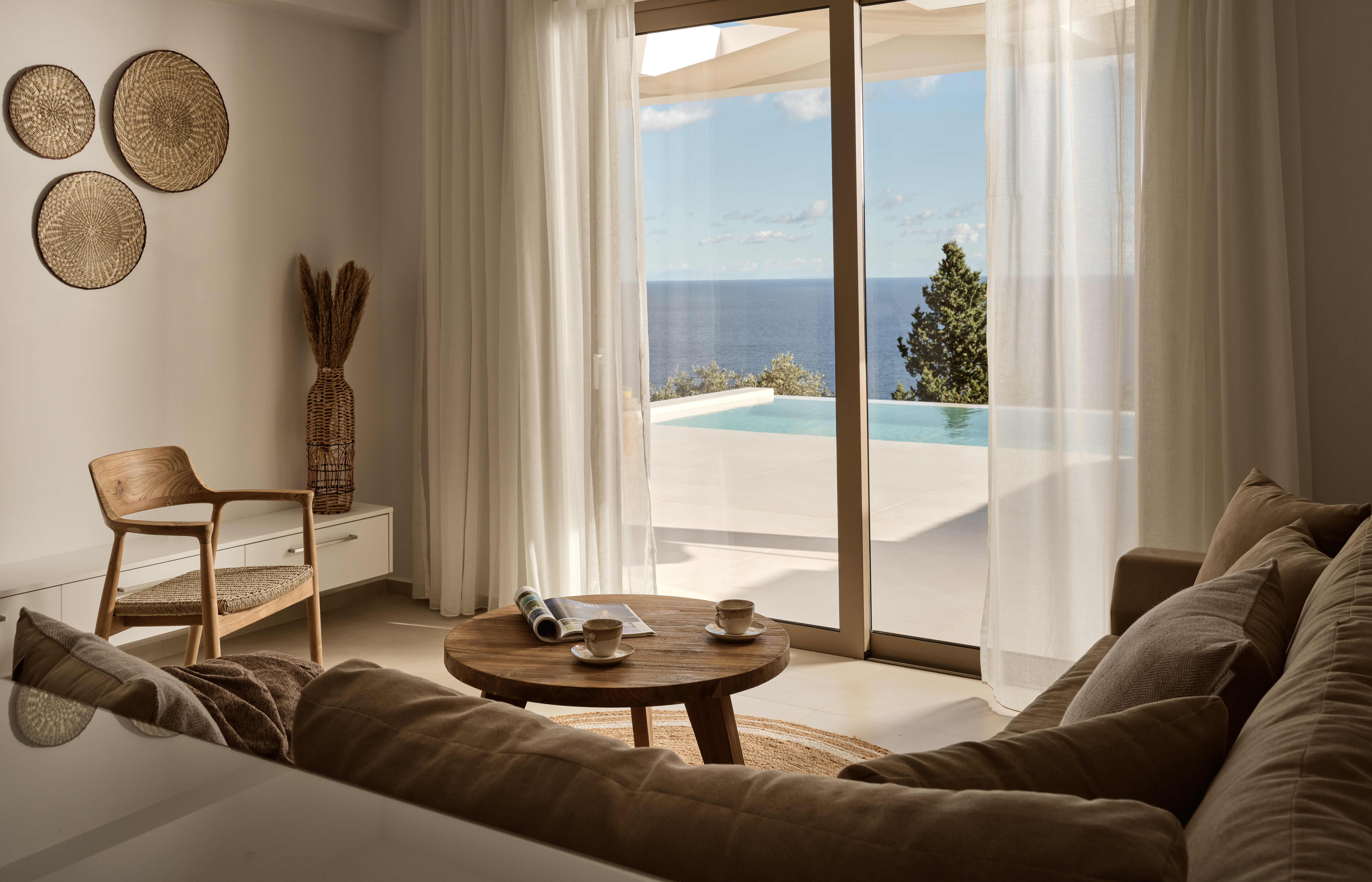 Contemporary living space with woven wall baskets, wood furniture, and floor-to-ceiling windows overlooking pool and sea.