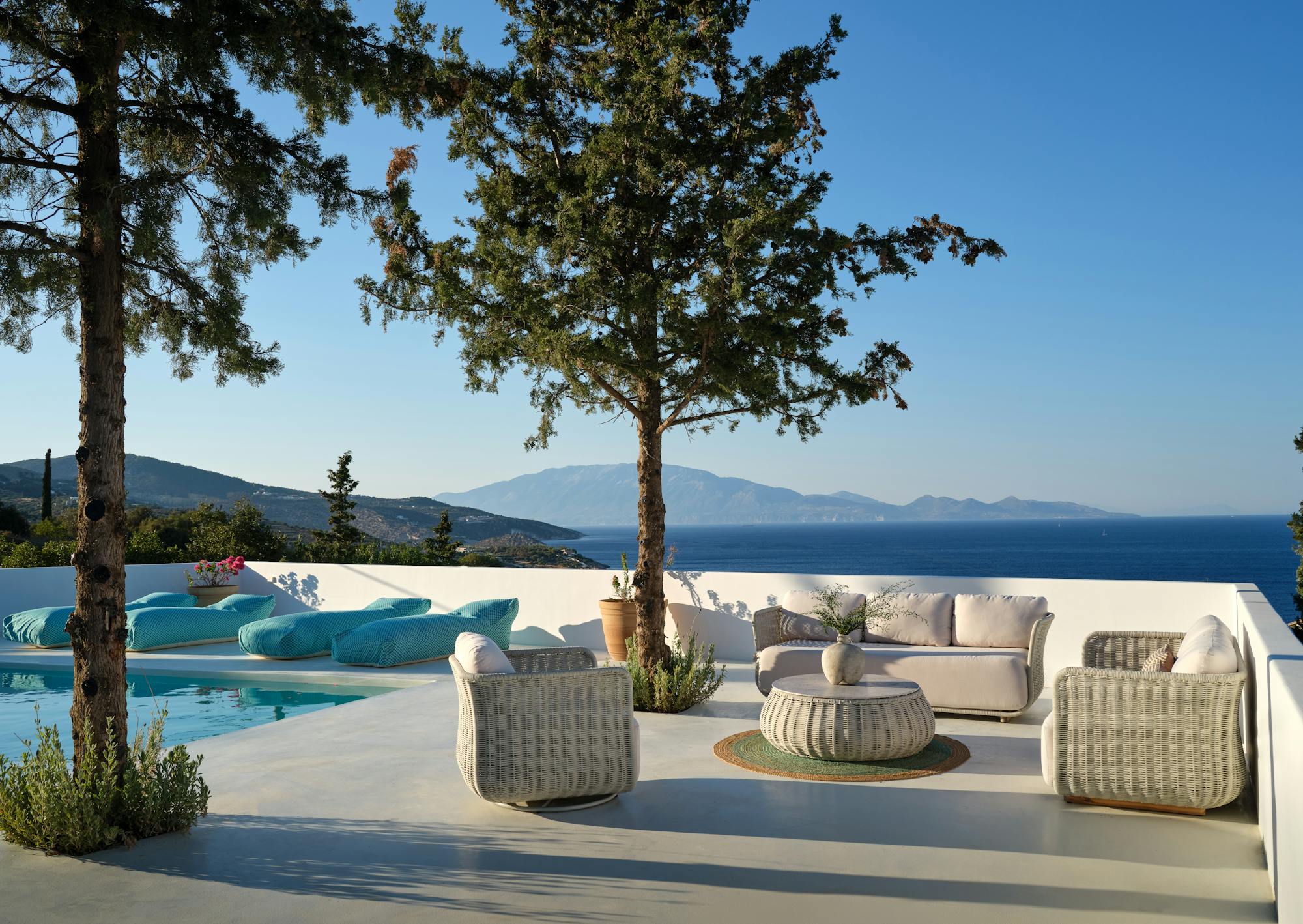 A white minimalist pool terrace features wicker furniture with white cushions under mature trees, overlooking distant islands and blue water.
