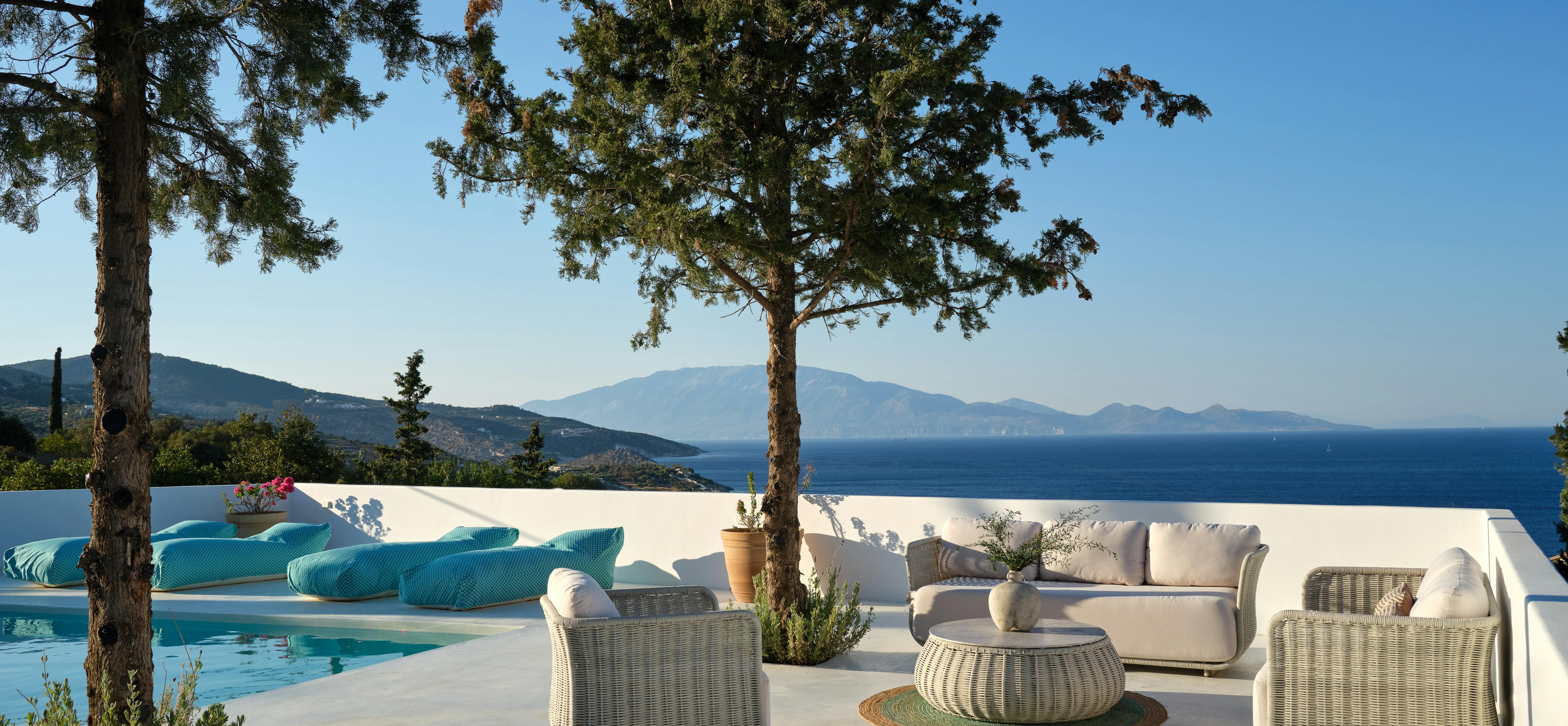 A white minimalist pool terrace features wicker furniture with white cushions under mature trees, overlooking distant islands and blue water.