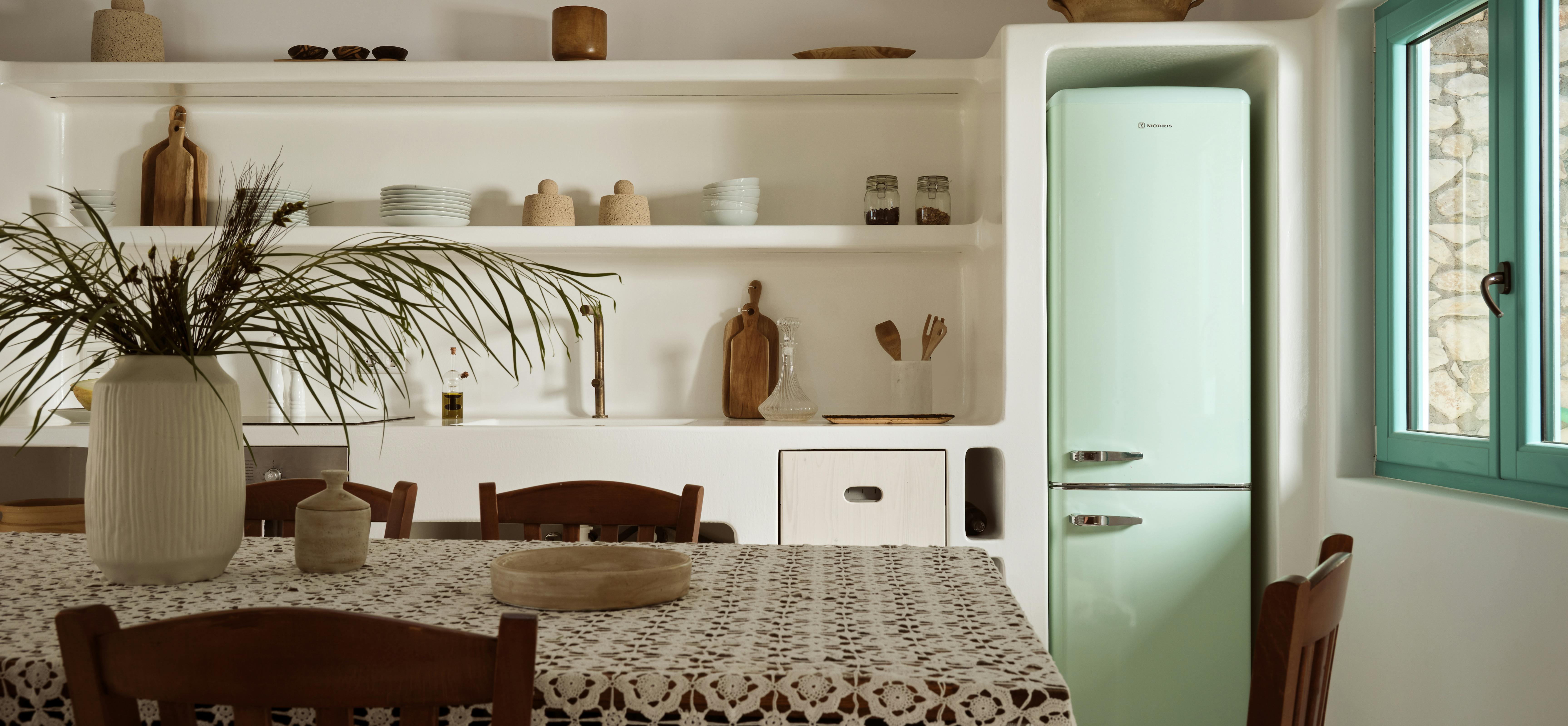 A white kitchen showcases open shelving with wooden bowls, lace-covered table, wooden chairs, and a mint green vintage refrigerator near turquoise windows.