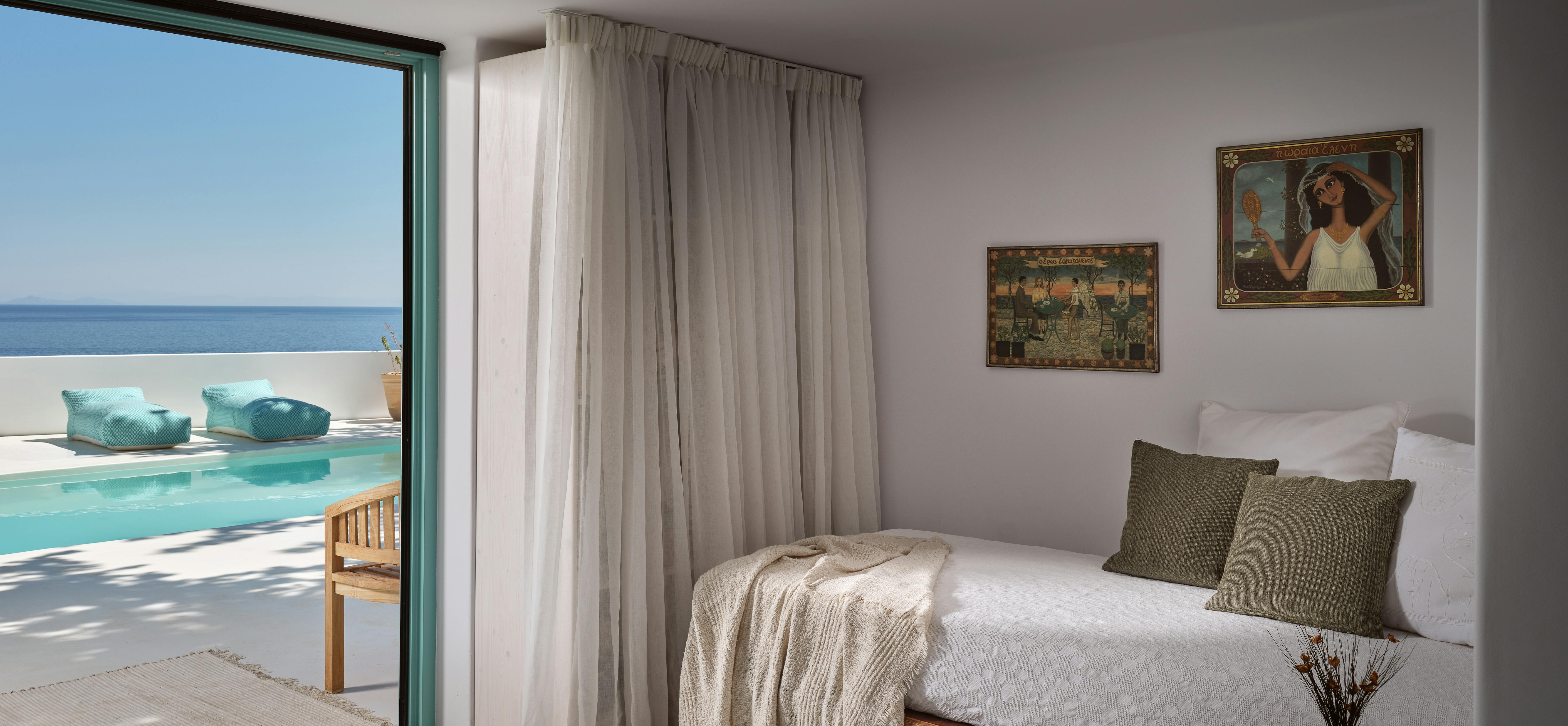 A serene bedroom with wood platform bed and vintage artwork opens directly to the pool terrace through floor-to-ceiling glass and flowing curtains.