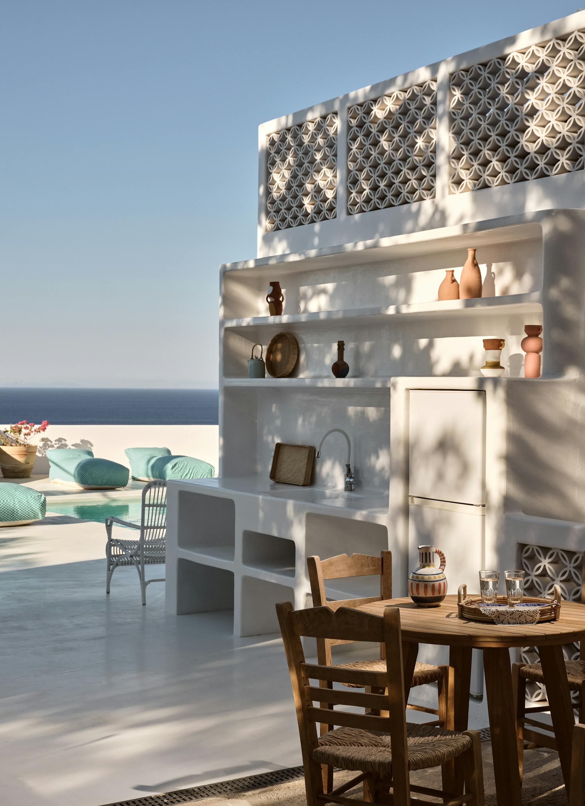A white outdoor kitchen area with built-in shelving displays pottery and connects to the pool terrace, featuring decorative lattice screens and wooden dining furniture.
