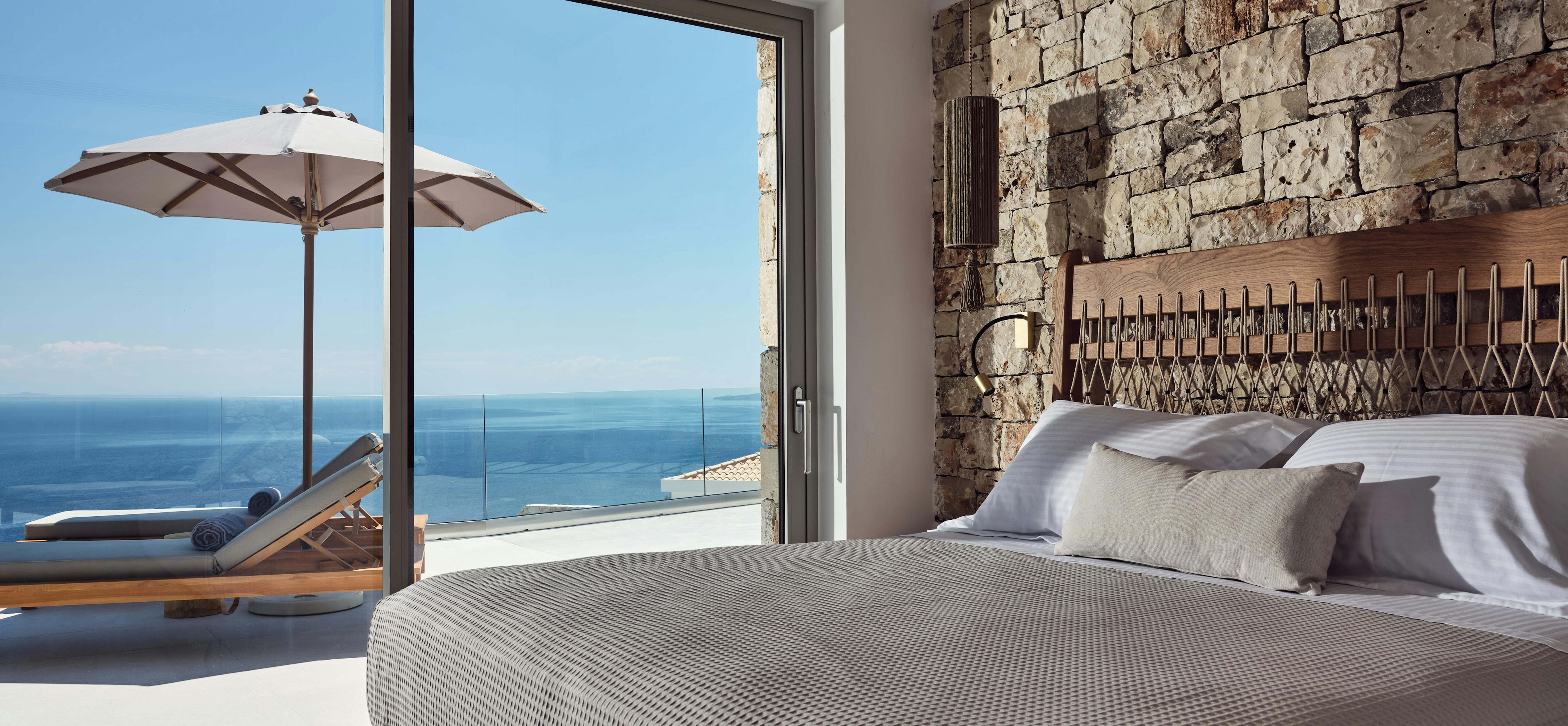 A luxurious bedroom with textured grey bedding opens through sliding glass doors to a terrace with wooden loungers, white umbrella, and endless ocean views beyond a rustic stone accent wall.