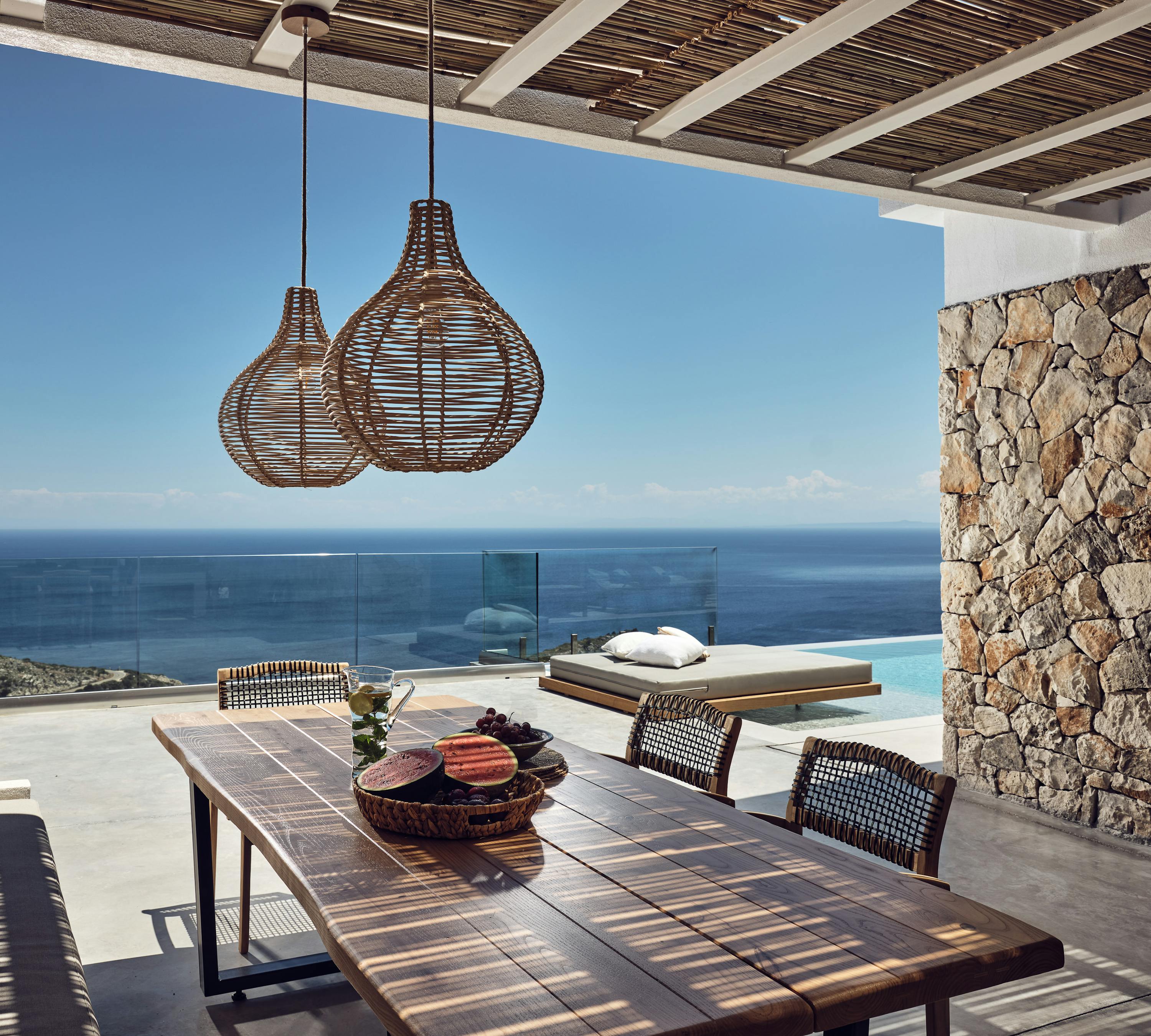 A covered dining terrace with wooden table and woven pendant lights frames views of an infinity pool, daybed, glass railings, and the sparkling sea beyond stone walls.