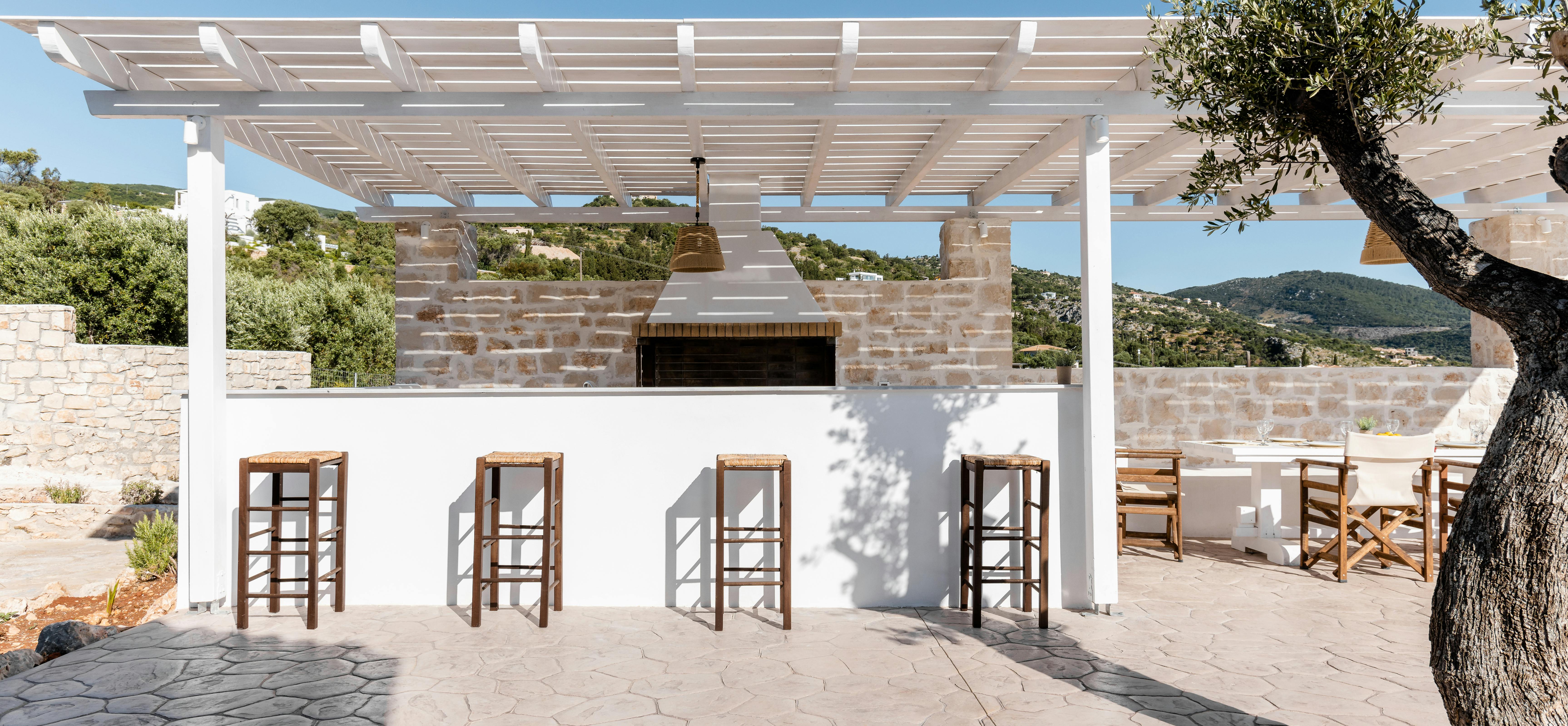 Outdoor bar area with four wooden stools under white pergola beside hexagonal paving and olive tree.