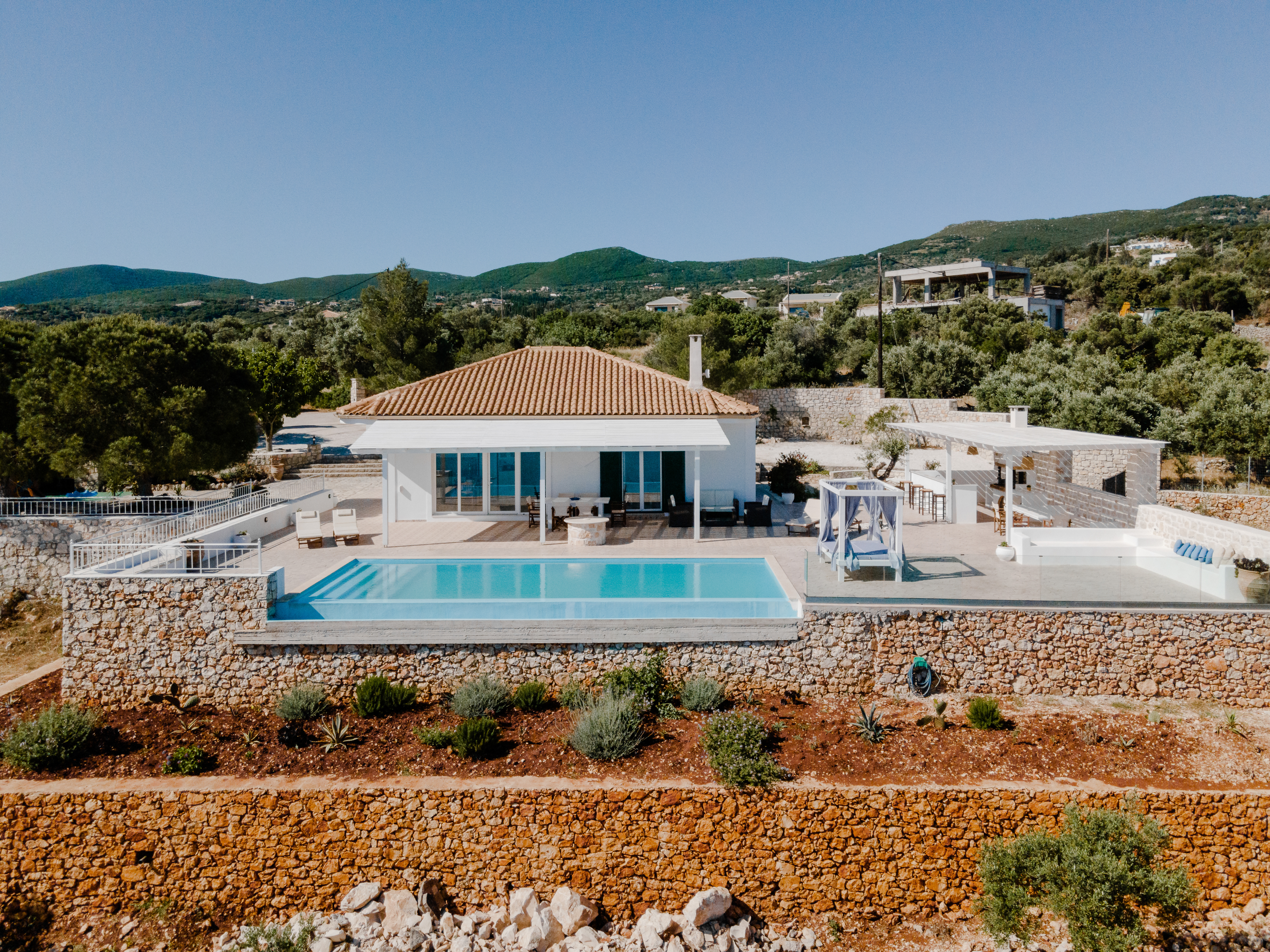 Aerial view of white Mediterranean villa with terracotta roof, infinity pool, and terraced stone walls overlooking mountainous landscape.