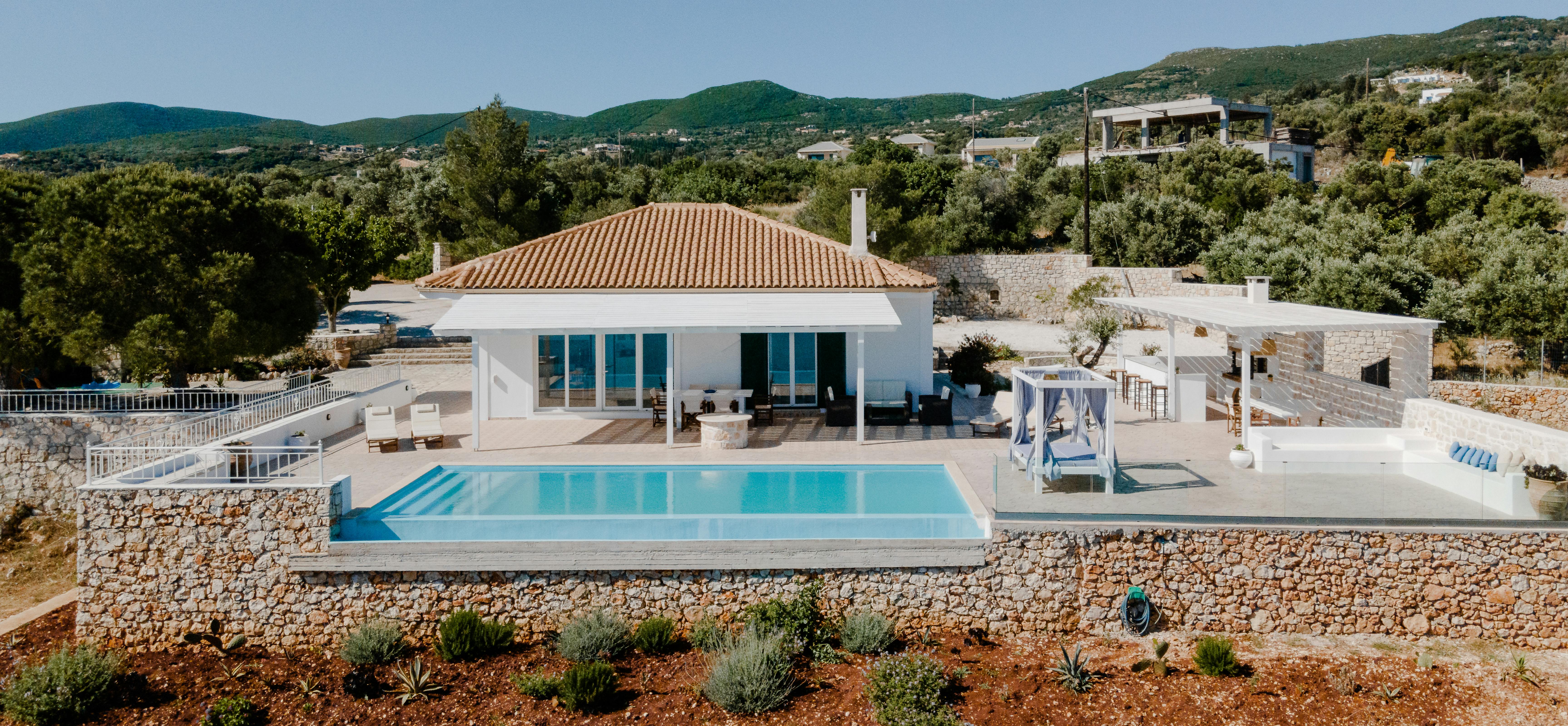 Aerial view of white Mediterranean villa with terracotta roof, infinity pool, and terraced stone walls overlooking mountainous landscape.