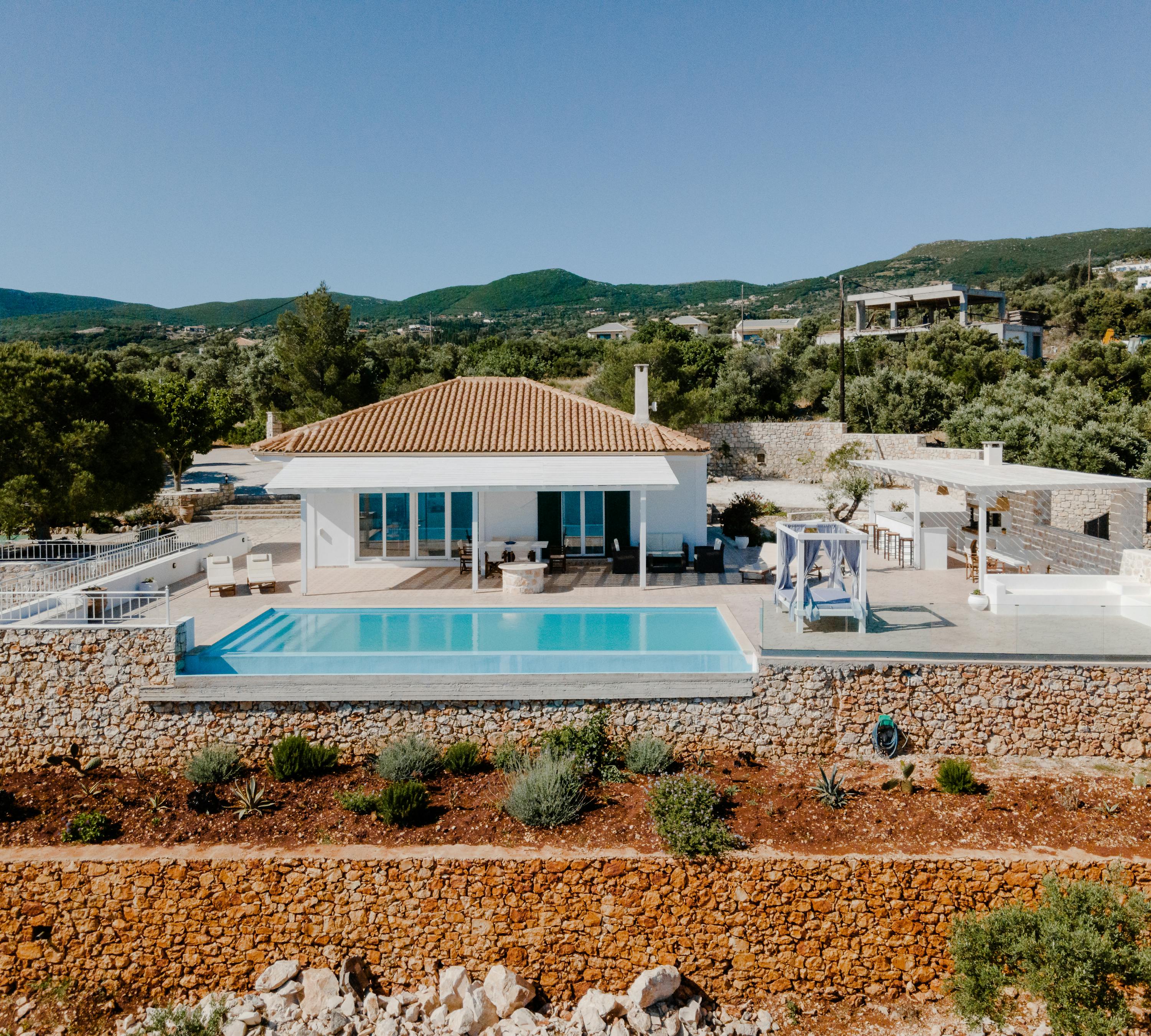 Aerial view of white Mediterranean villa with terracotta roof, infinity pool, and terraced stone walls overlooking mountainous landscape.